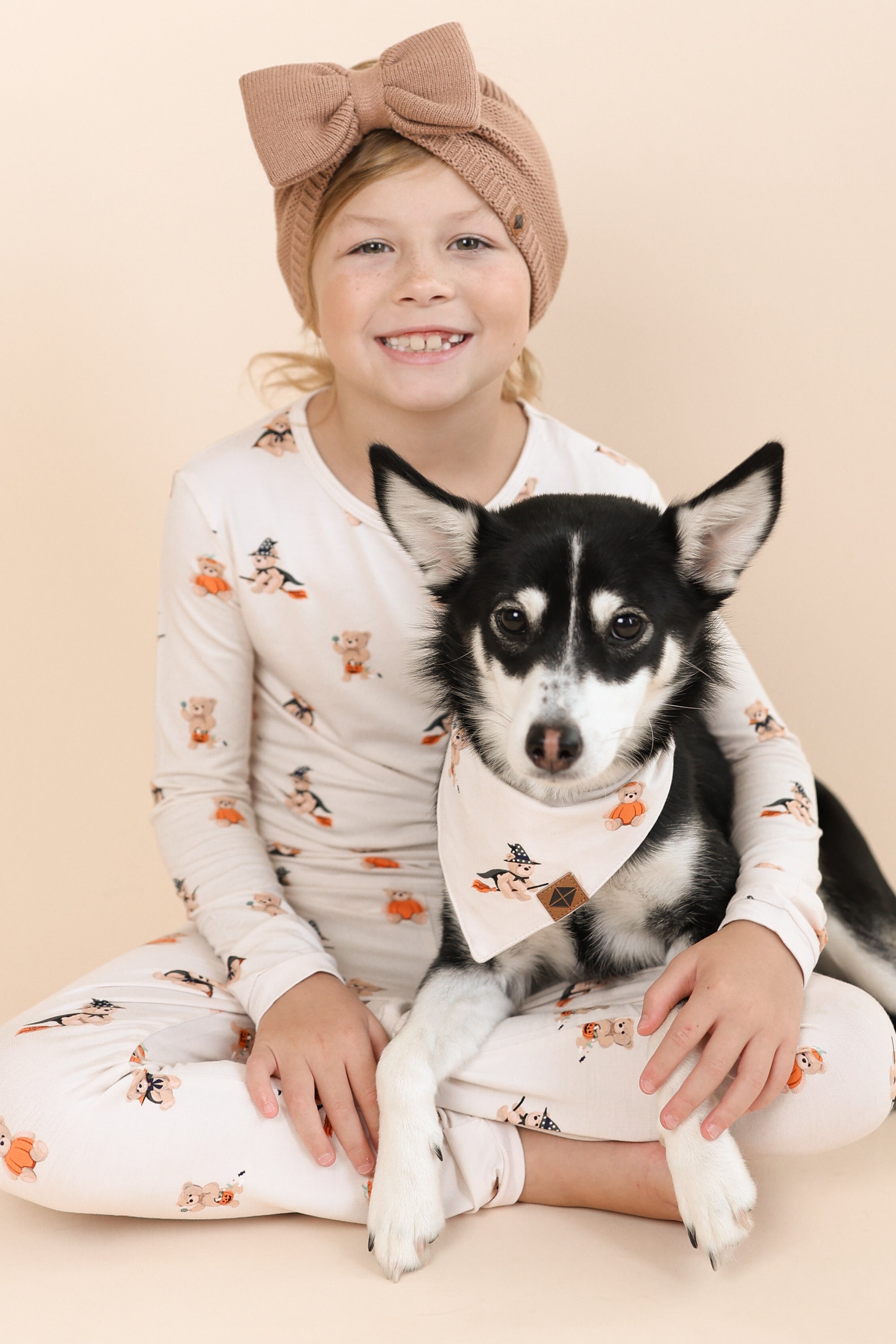Black and white Dog wearing Dog Bandana in Halloween Teddy print sitting with a girl wearing a similar print long sleeve pajama set and a latte headwrap on a beige background