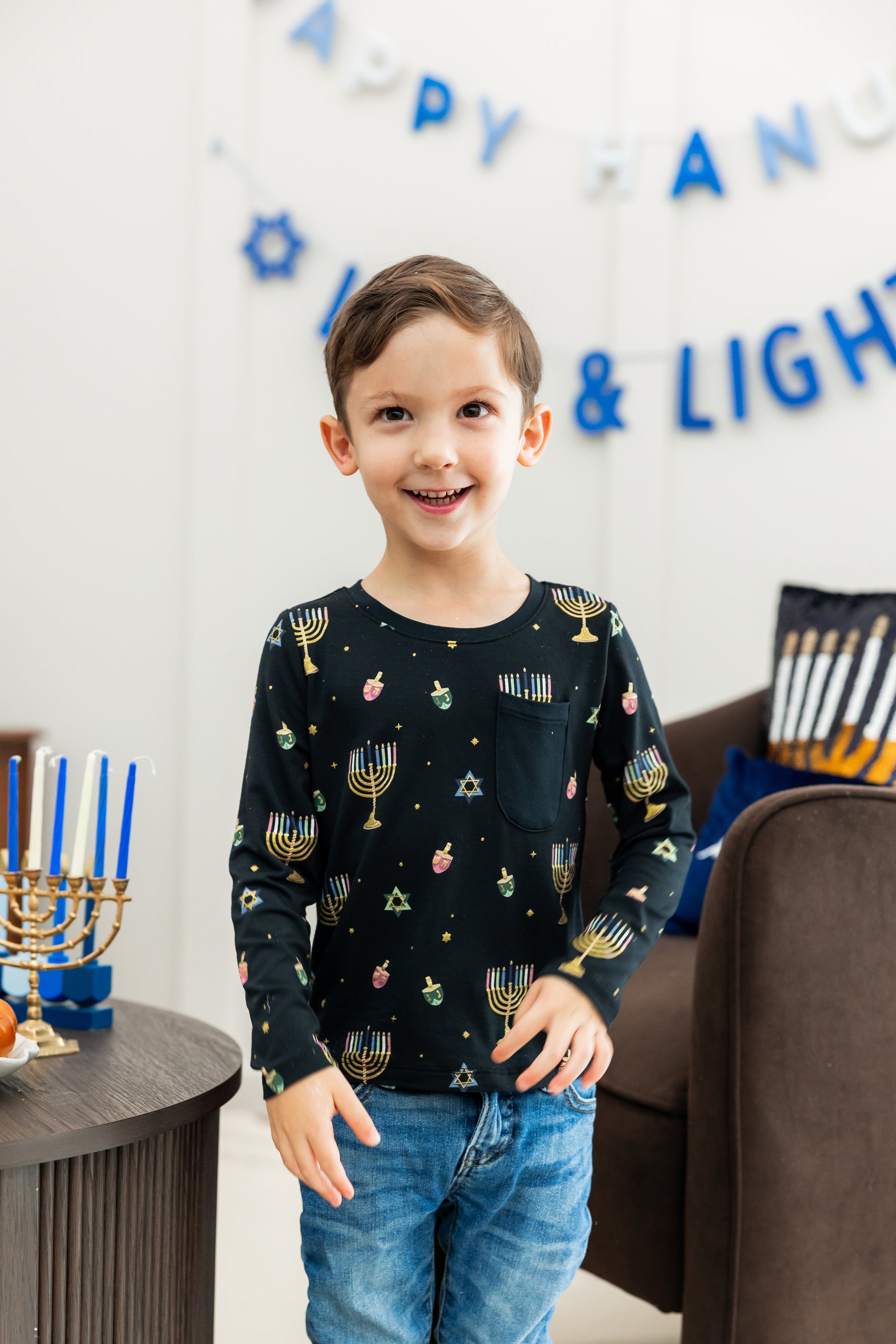 Toddler smiling wearing a long sleeve crew neck tee in hnukkah and blue jeans
