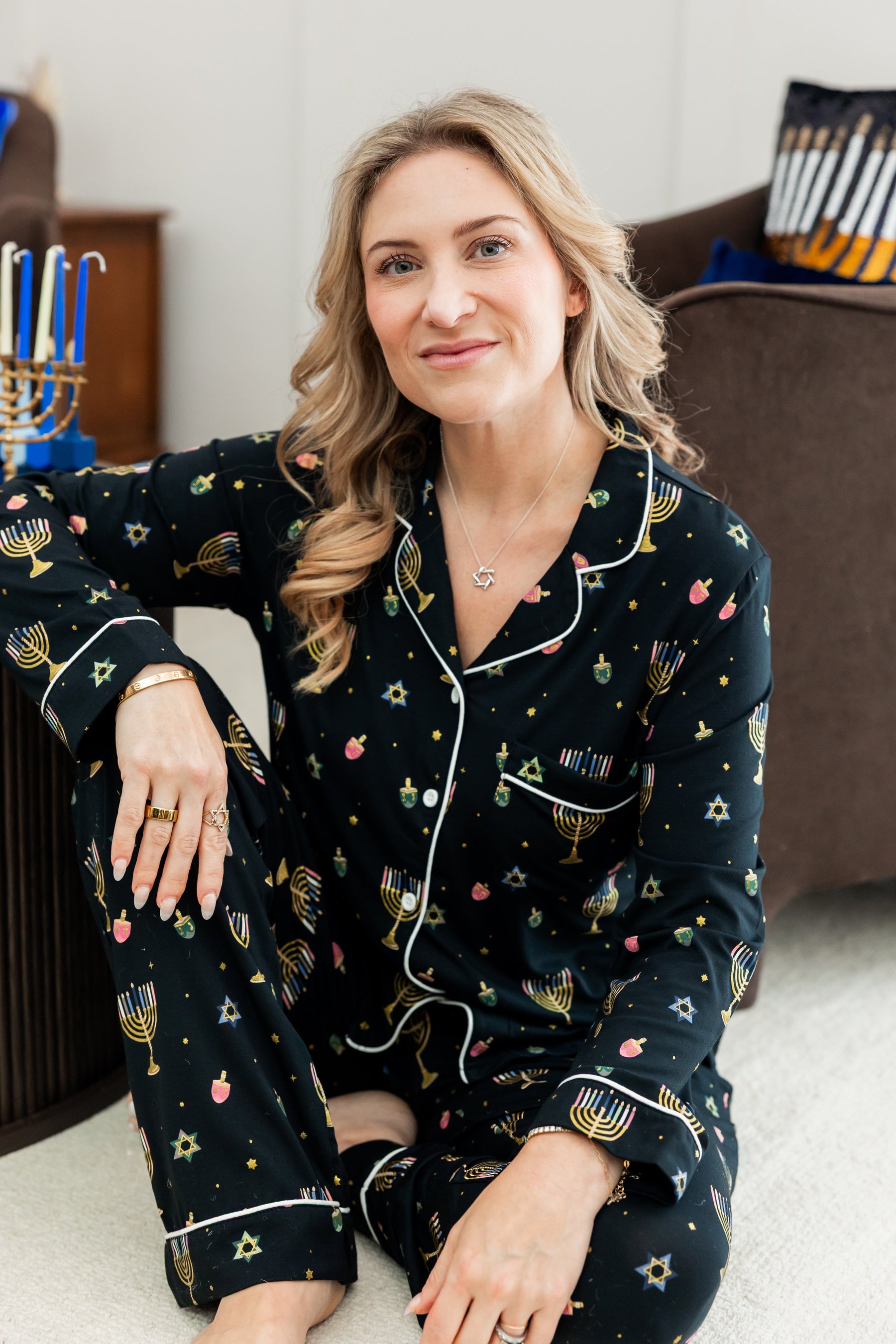 Woman sitting on the ground with one leg bent in and the other propped up with her arm resting on it smiling wearing long sleeve pajamas in hanukkah print