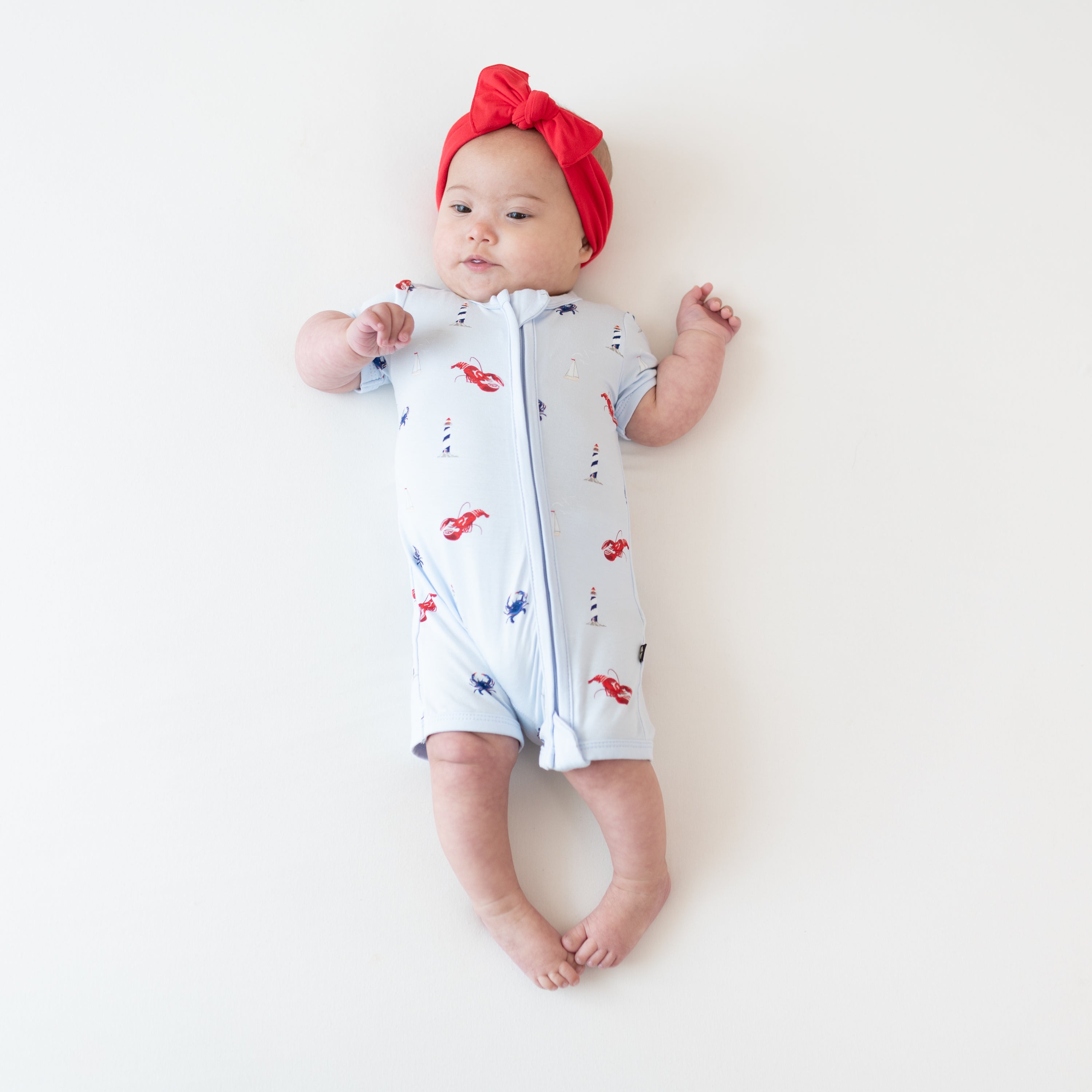 Infant girl laying on a blanket wearing the Zipper Shortall Romper in Harbor paired with a Knotted Bow Headband in Cardinal