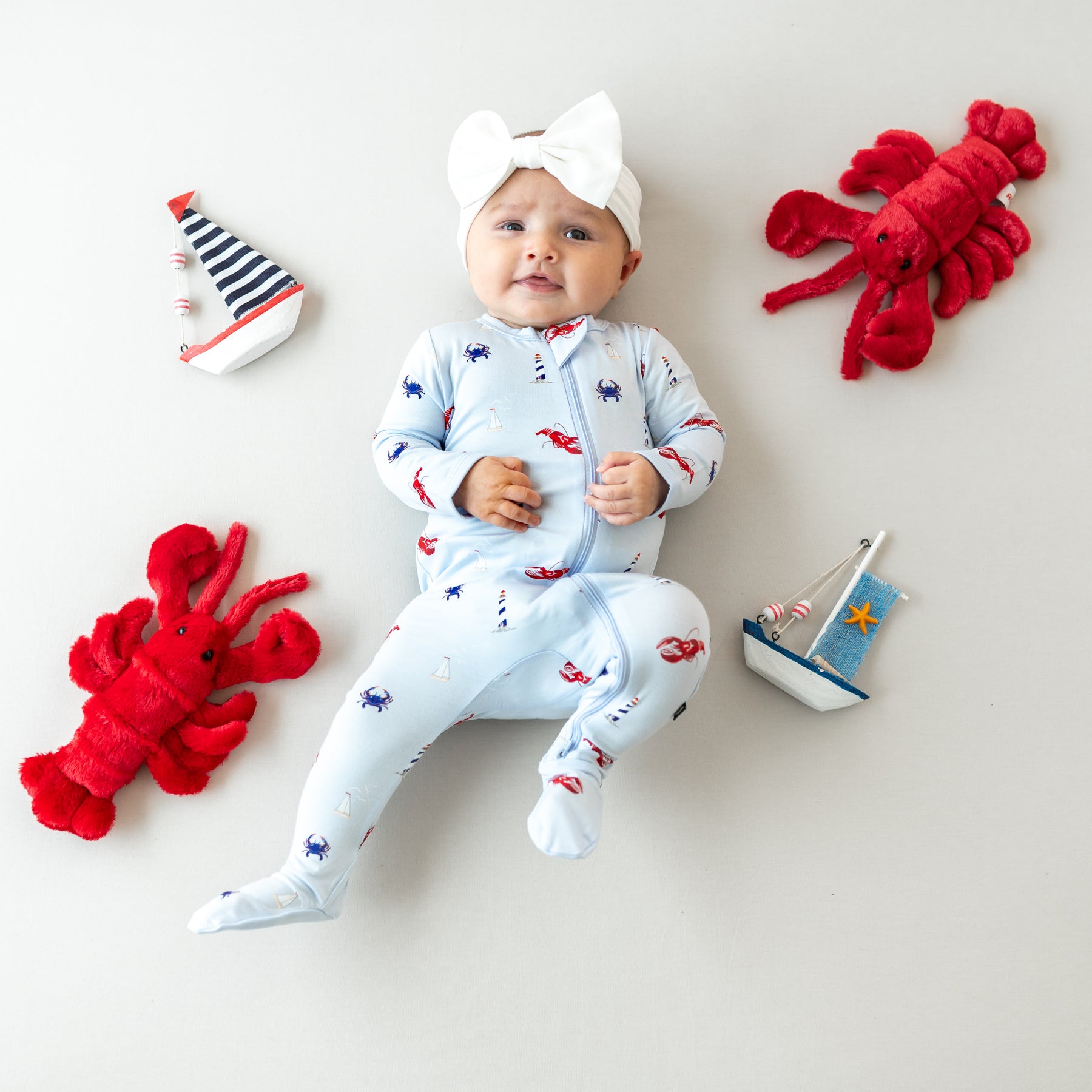 Smiling infant laying down with plush nautical themed toys around her wearing the Zippered Footie in Harbor and Bow Headband in Cloud