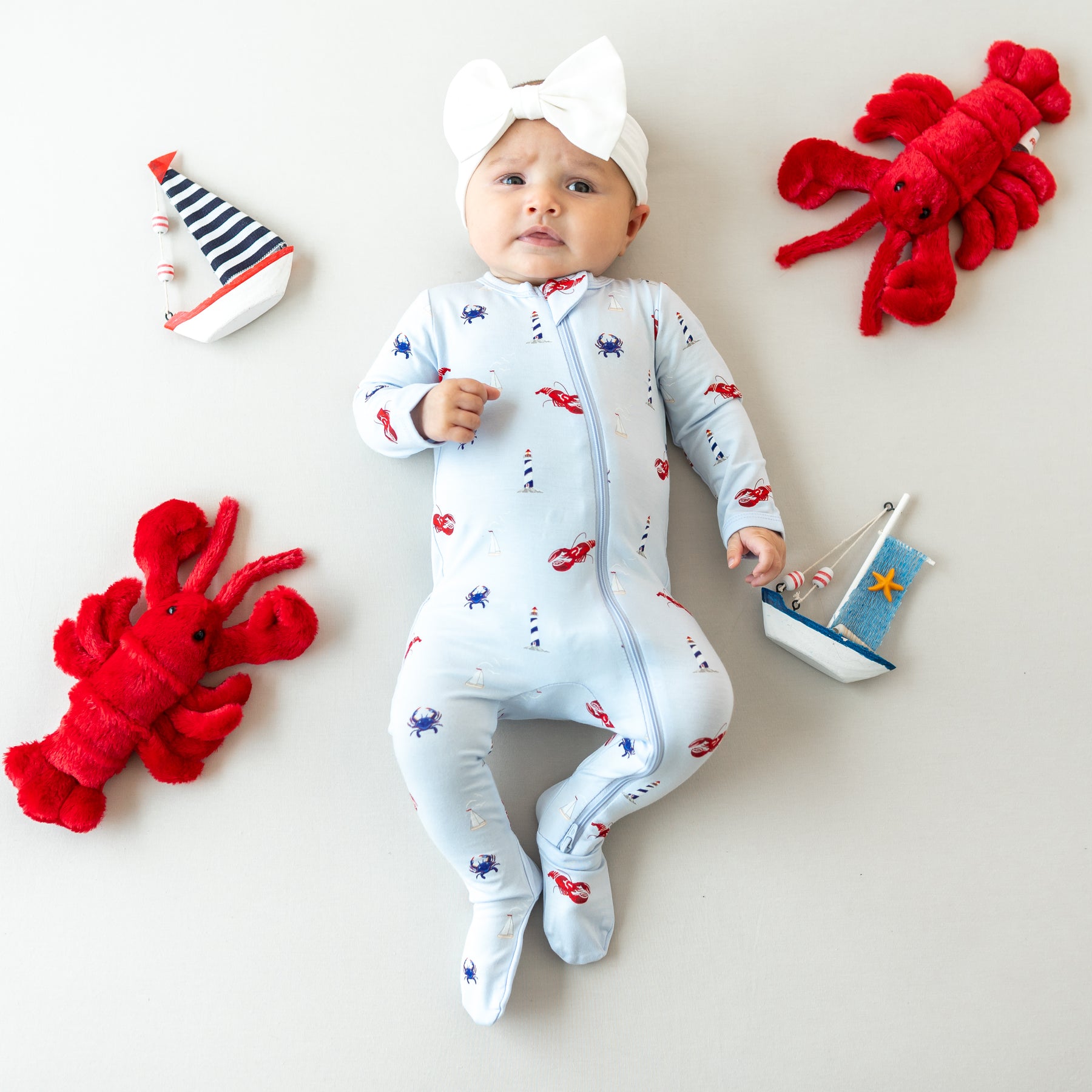 Infant girl laying down with plush nautical toys around her wearing the Zippered Footie in Harbor and the Bow Headband in Cloud