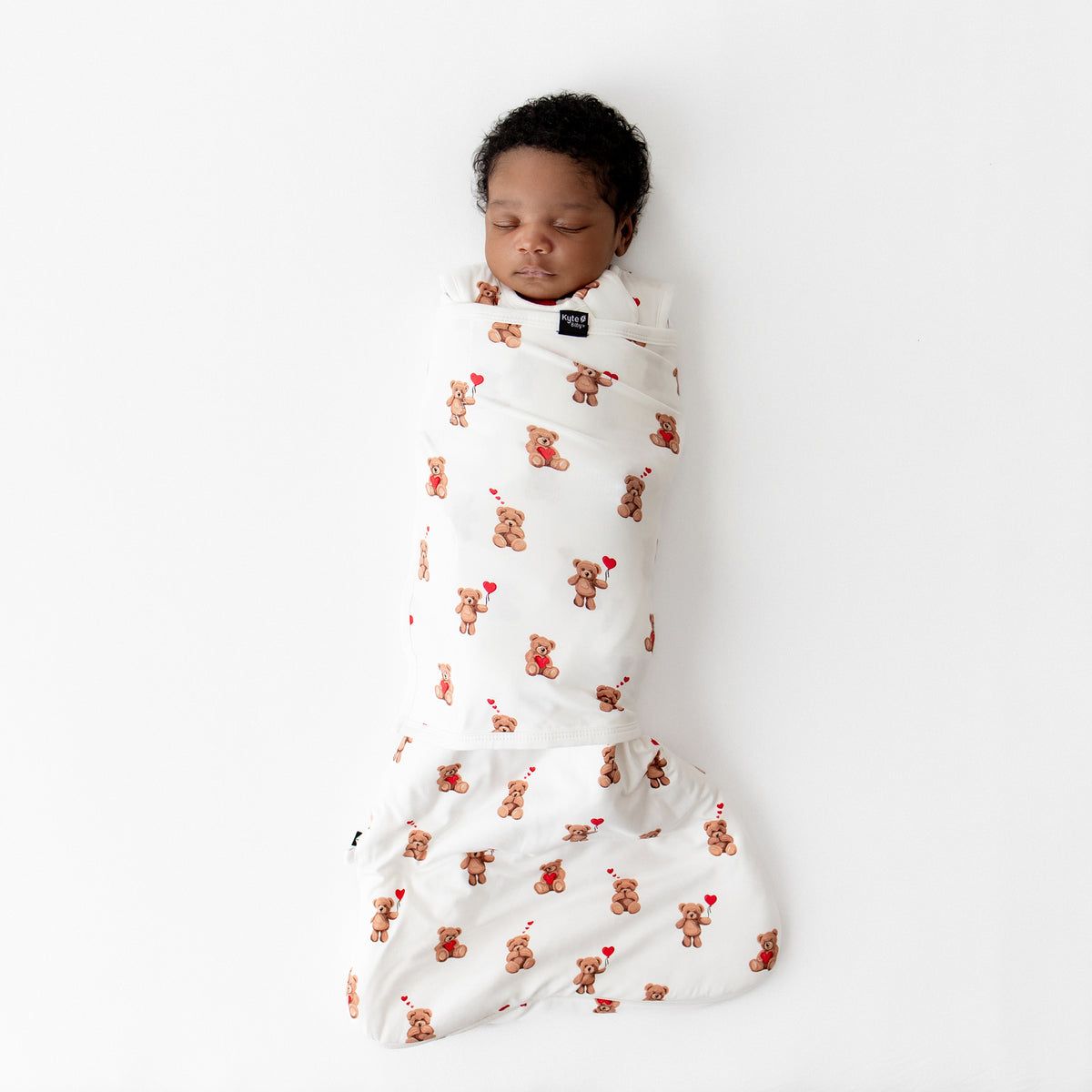 Sleeping newborn laying on a white surface swaddled in the soft and stretchy Sleep Bag Swaddler in Bear Hearts 1.0