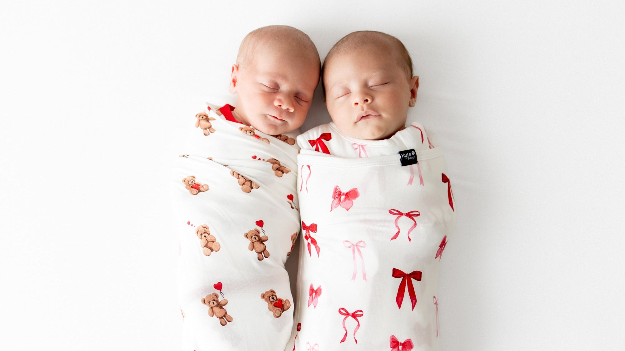Two babies wrapped in Valentines patterned swaddles on a white background