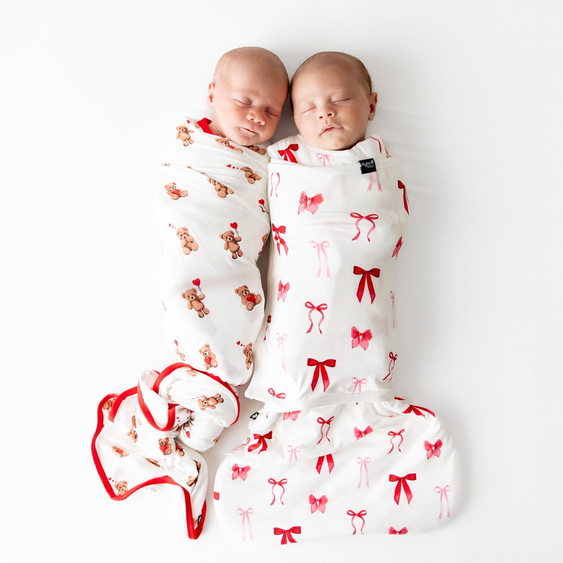 Two sleeping newborns laying side by side. One newborn wearing the Swaddle Blanket in Bear Hearts and the other wearing the sleep bag swaddler in small love bow