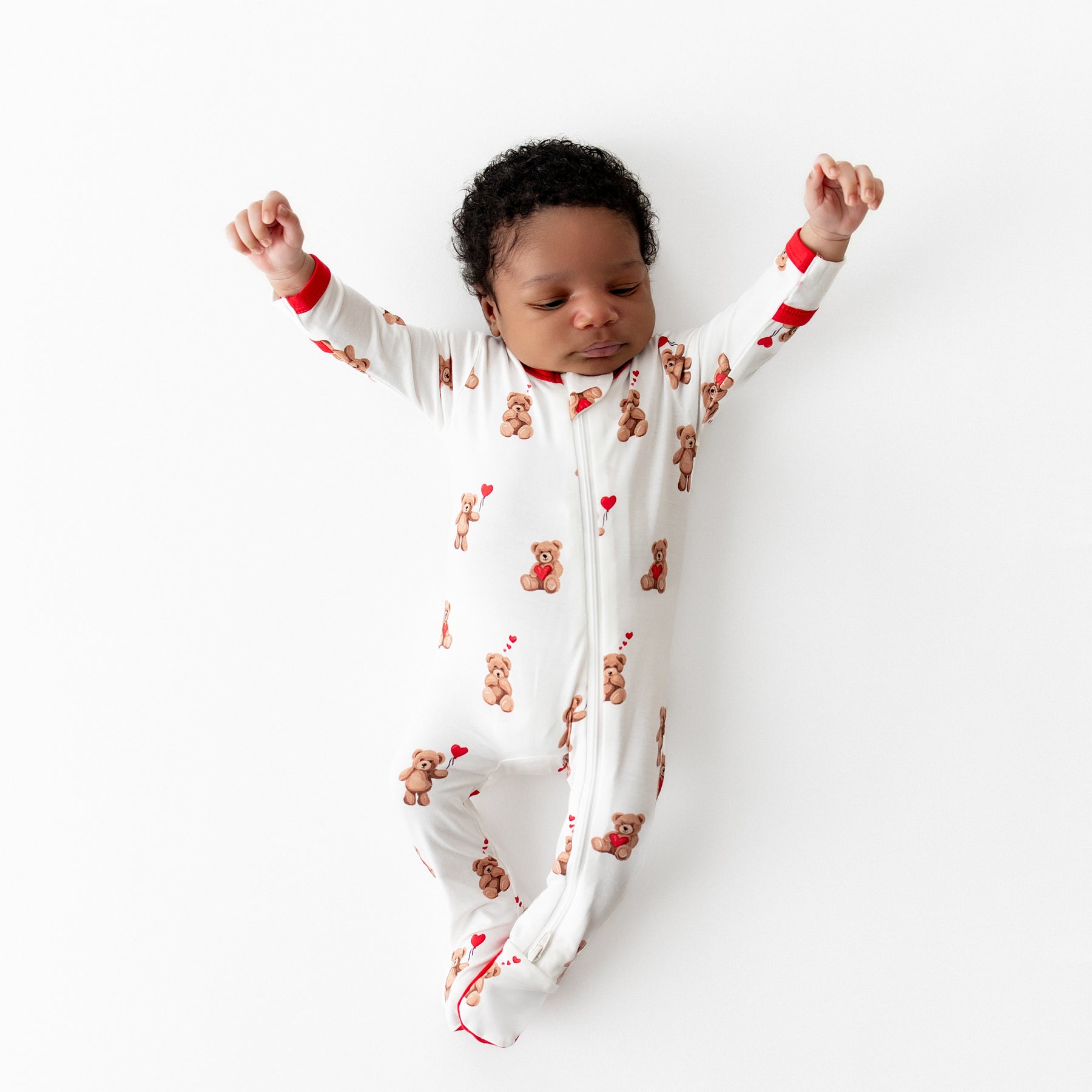 Stretching newborn laying on a white surface wearing the breathable Zippered Footie in Bear Hearts