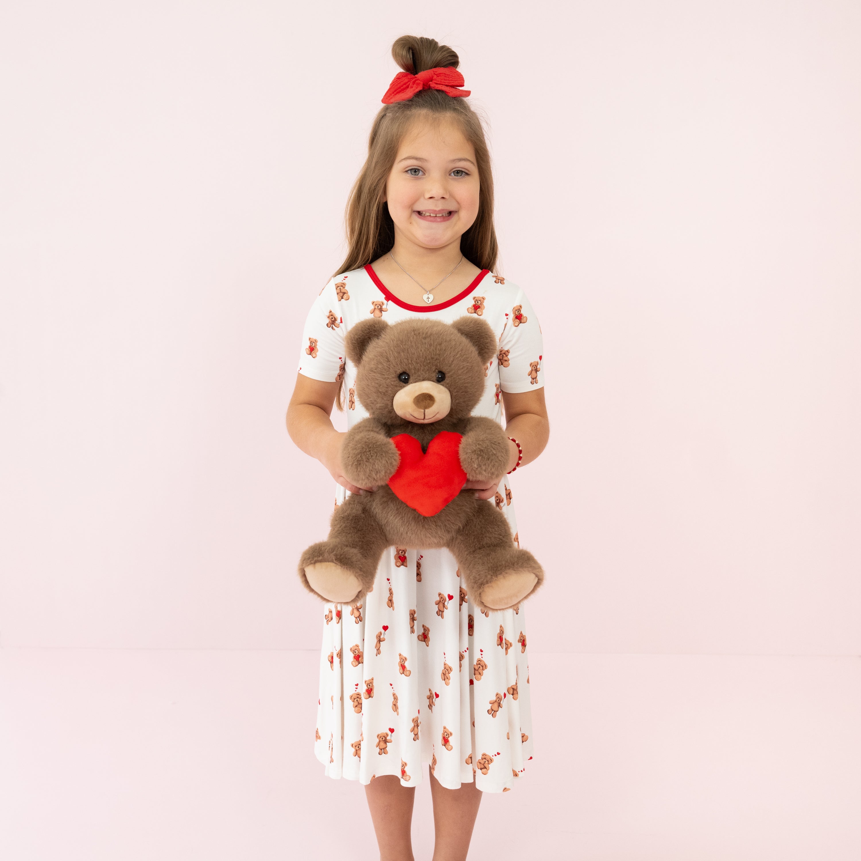 Young smiling girl standing in front of a pink background wearing the Twirl Dress in Bear Hearts holding Kyte Baby's Valentine's Teddy Bear