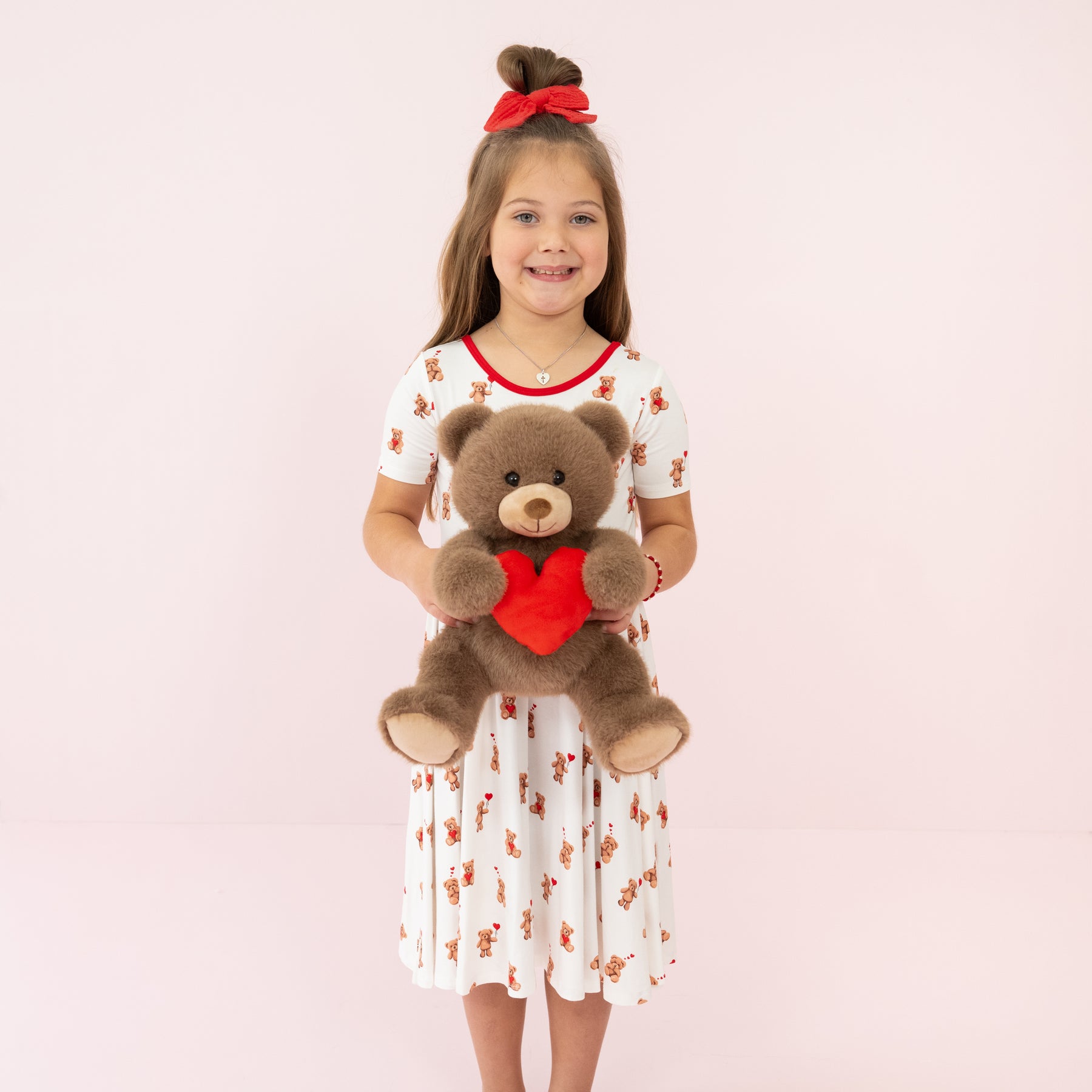 Young smiling girl standing in front of a pink background wearing the Twirl Dress in Bear Hearts holding Kyte Baby's Valentine's Teddy Bear