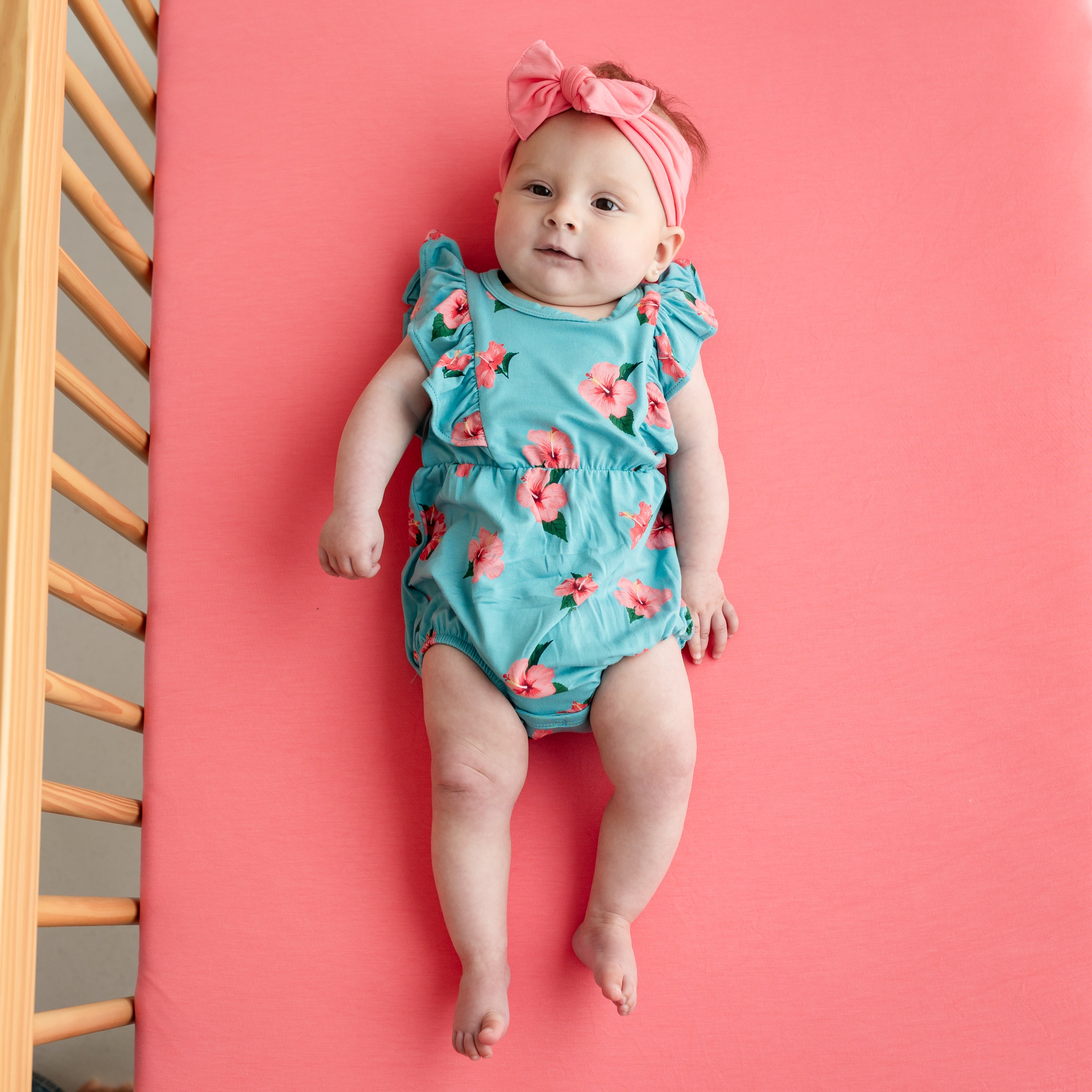 Baby laying wearing Kyte Baby Bubble Romper in Hibiscus on guava crib sheet