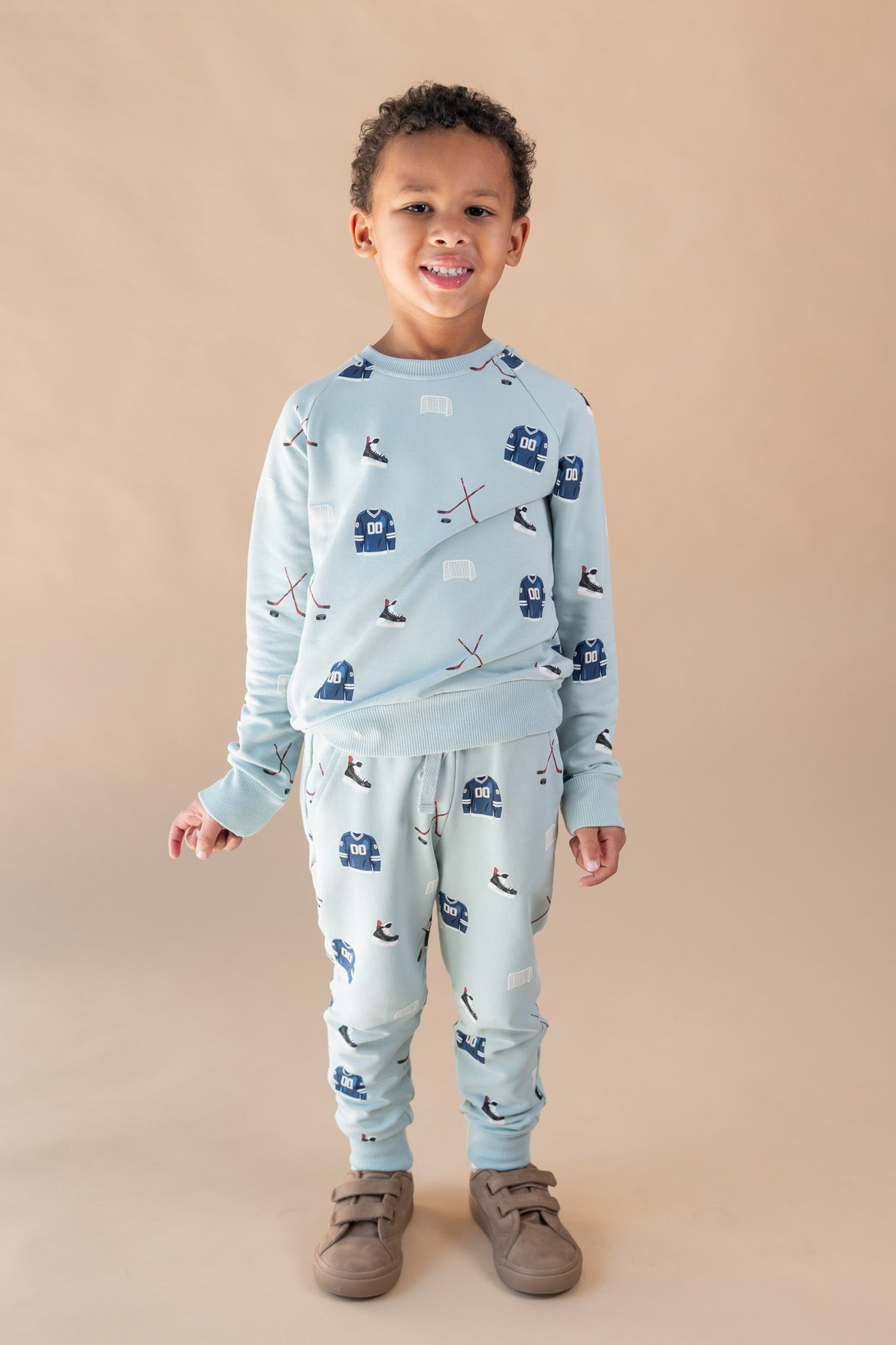 A young child wearing a light blue long-sleeve set with a dark blue and white ski pattern, paired with brown shoes, standing against a solid beige background.