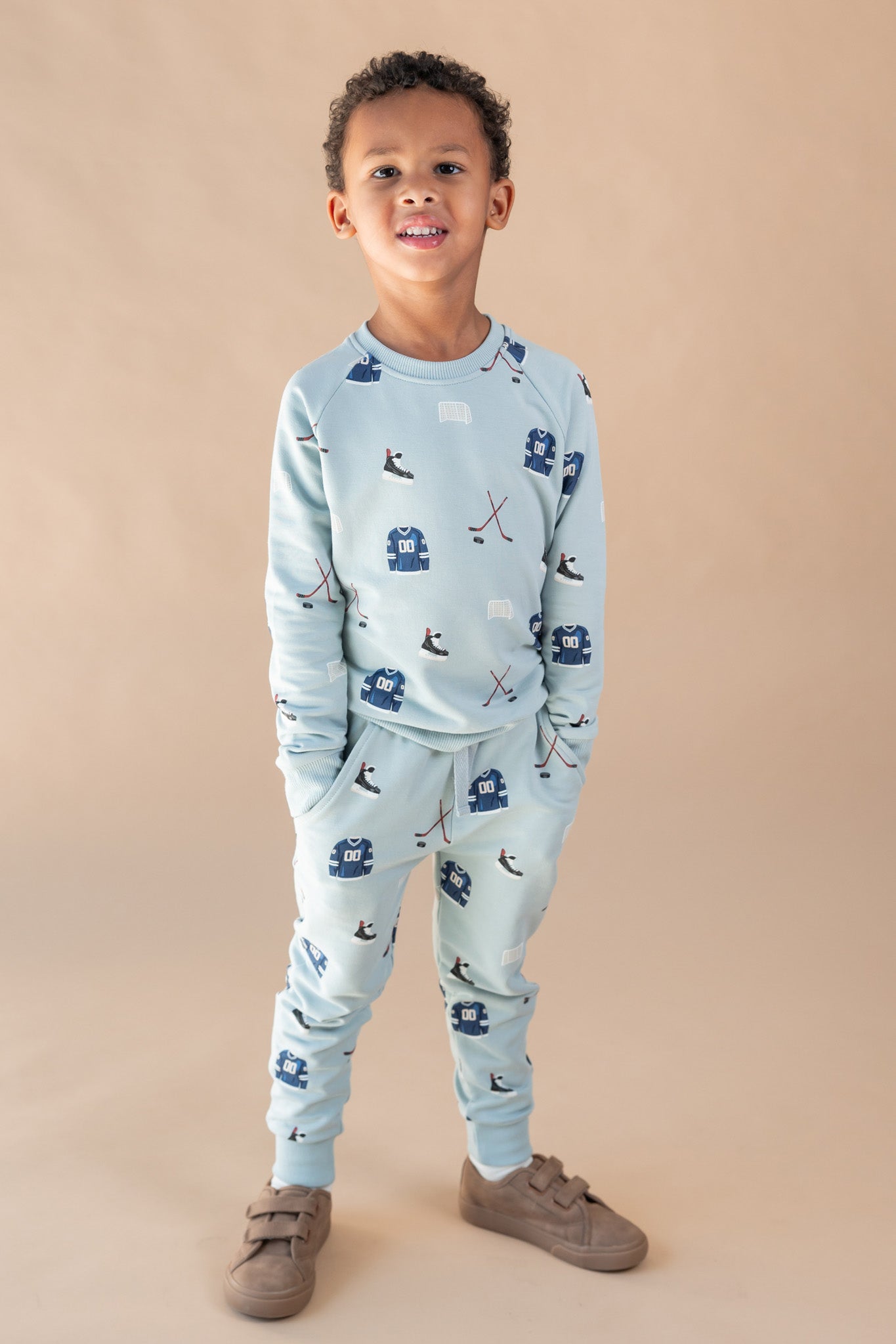 A young boy modeling a light blue two-piece set with a hockey print, consisting of a long-sleeve top and matching jogger pants, paired with brown shoes, against a solid beige studio background.