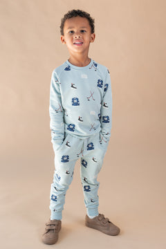 A young boy modeling a light blue two-piece set with a hockey print, consisting of a long-sleeve top and matching jogger pants, paired with brown shoes, against a solid beige studio background.