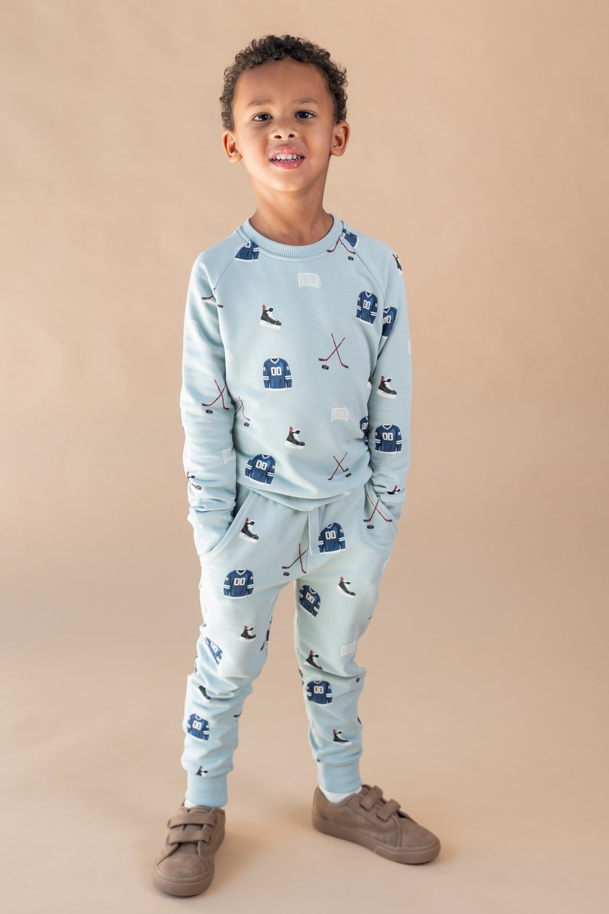 A young boy modeling a light blue two-piece set with a hockey print, consisting of a long-sleeve top and matching jogger pants, paired with brown shoes, against a solid beige studio background.