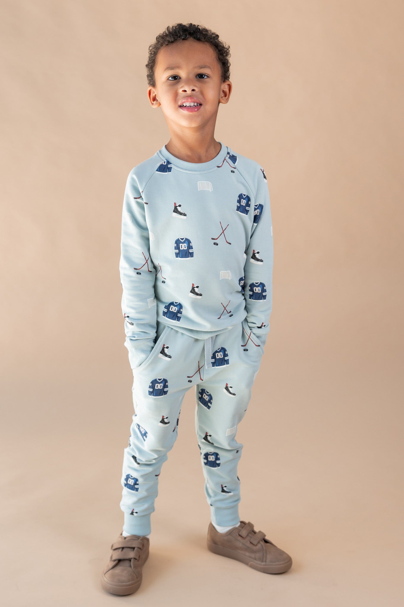 A young boy modeling a light blue two-piece set with a hockey print, consisting of a long-sleeve top and matching jogger pants, paired with brown shoes, against a solid beige studio background.