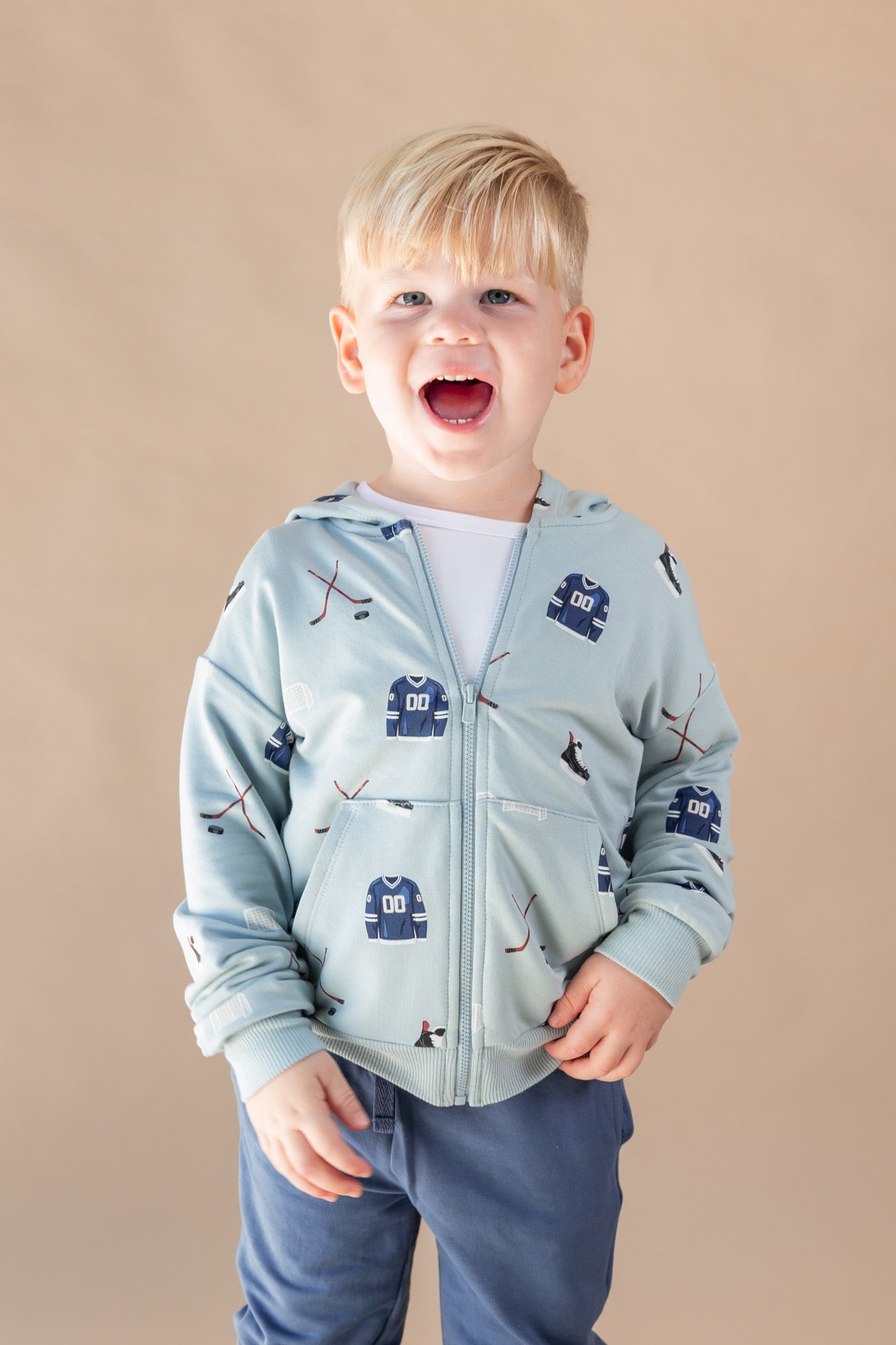 A young blonde boy wearing a light blue zip-up hoodie with a hockey print, standing against a solid beige background.
