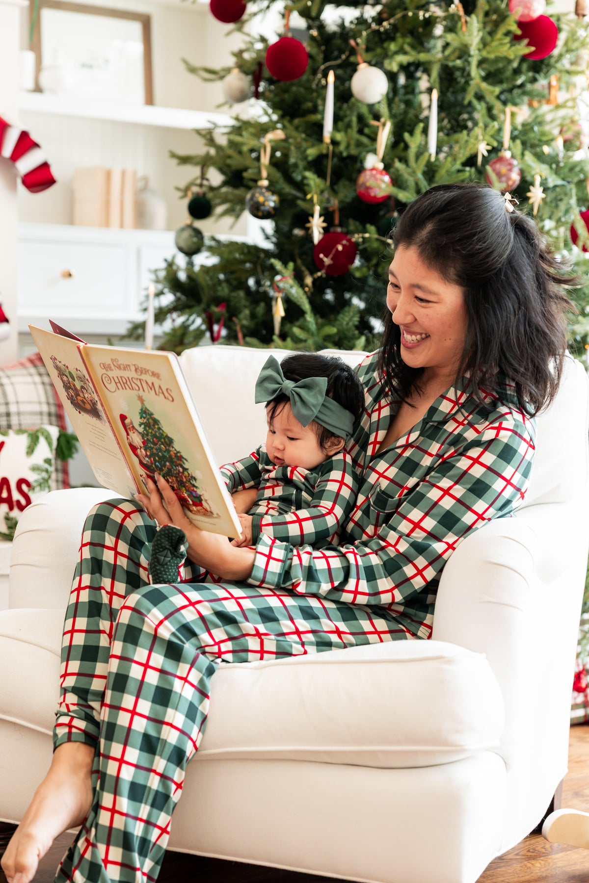 A woman and a child wearing matching green, red, and white plaid Christmas pajamas sit together on a white armchair in front of a decorated Christmas tree, reading a book.