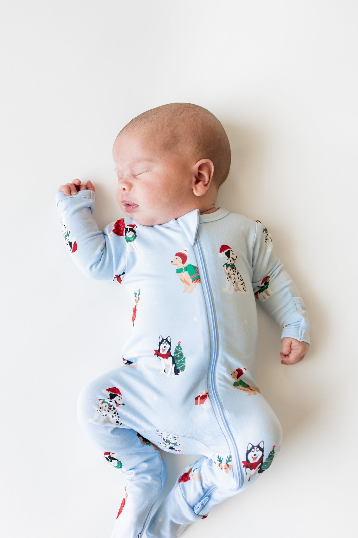A sleeping newborn baby wearing a light blue footed onesie with a Christmas print, lying on a plain white background.