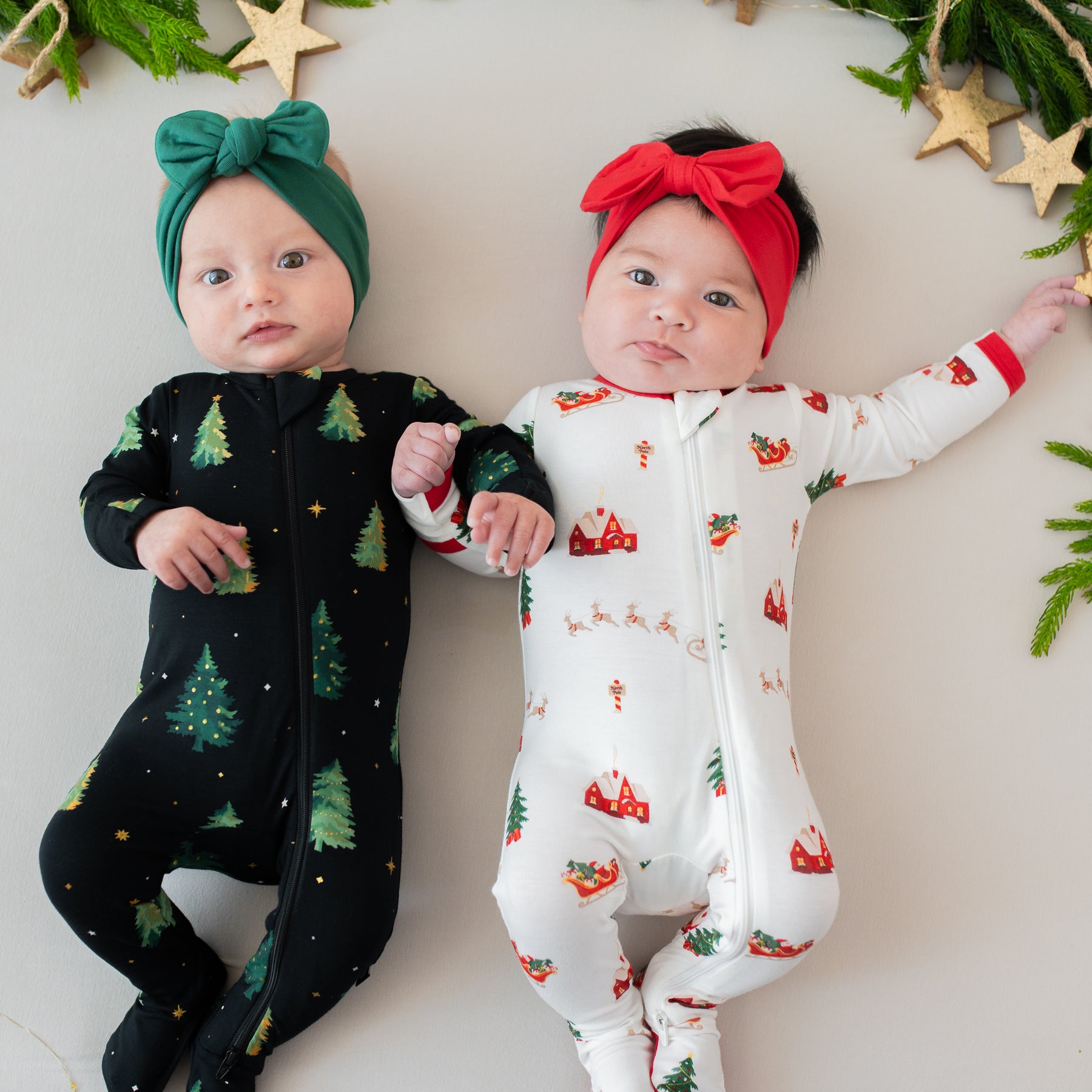 Two infants laying side by side with their arms intertwined wearing zippered footies in Santa Sleigh and twinkle trees with Bow Headband in Cardinal and Emerald