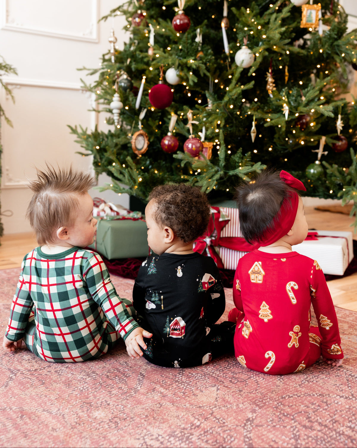 3 babies sitting by a christmas tree wearing festive holiday printed pajamas