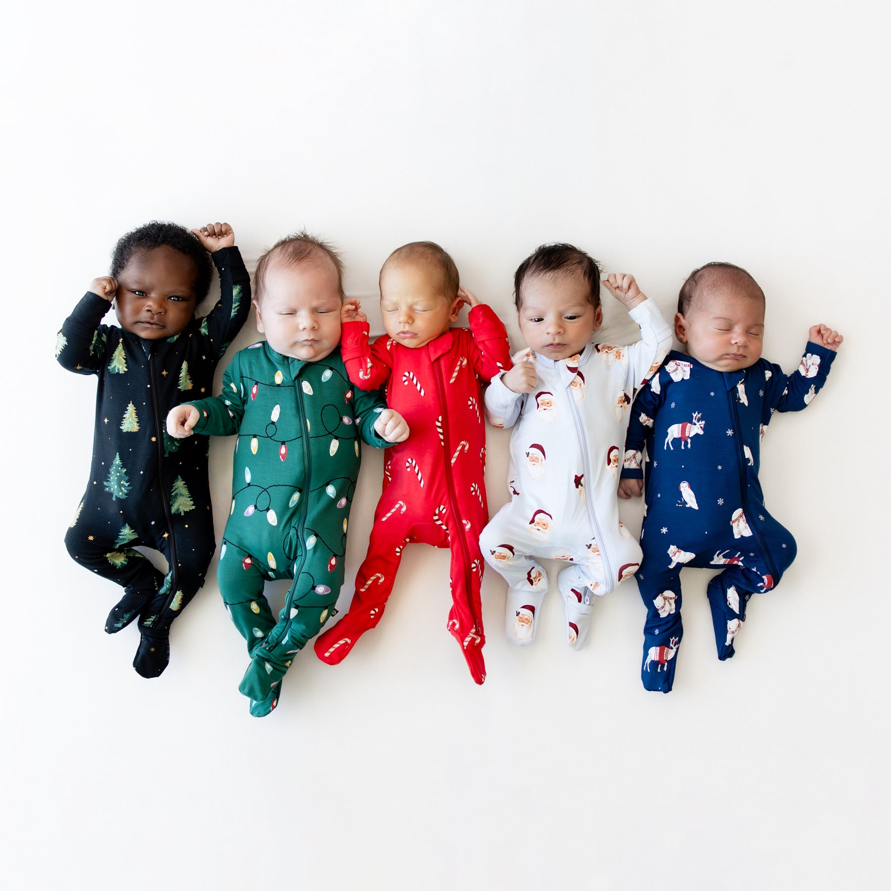 Five infant models laying side by side wearing Zippered Footie in Candy Cane, Vintage Santa, Frosty Friends, Merry and Bright and Twinkle Tree