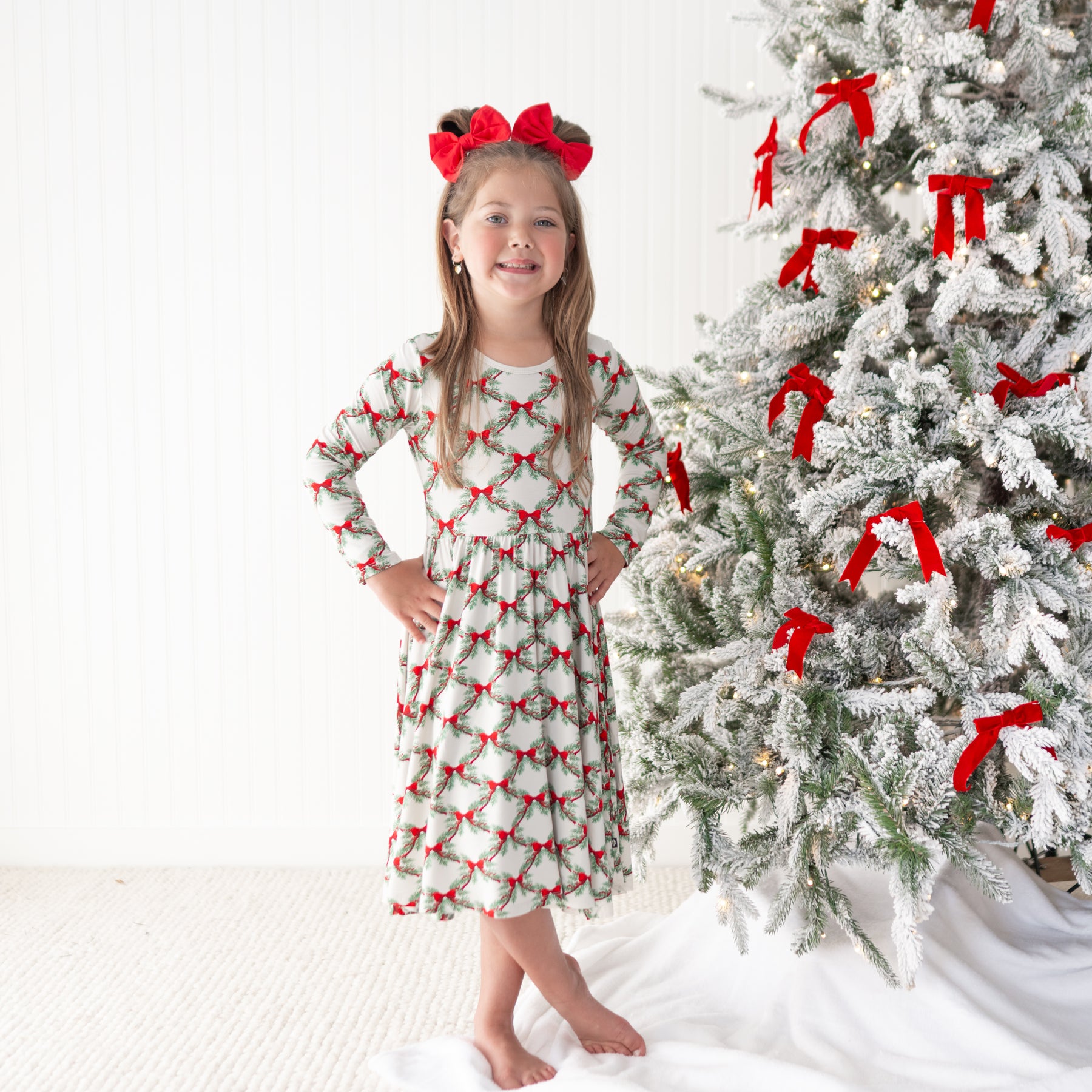 Young girl standing beside a decorated Christmas tree with her hands on her hips wearing the Long Sleeve Twirl Dress in Holiday Bow with red bows in her hair