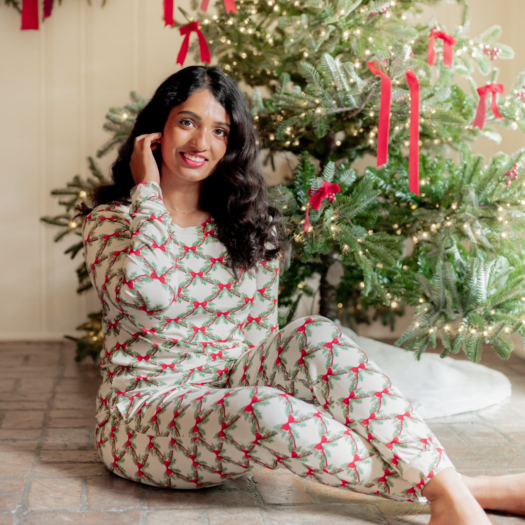 Smiling female model sitting on the floor wearing the Women's Jogger Pajama Set in Holiday Bow in front of a decorated Christmas Tree