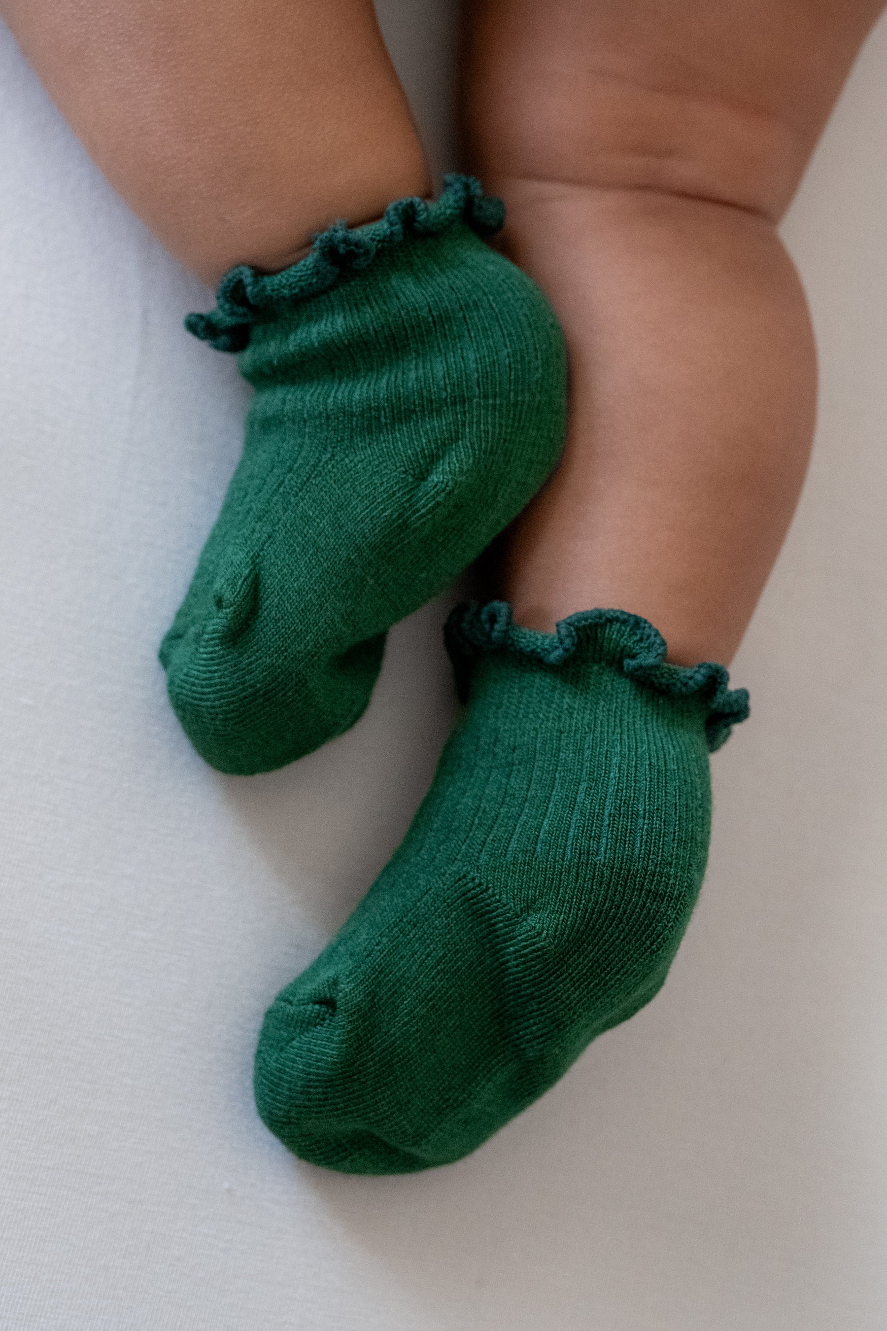 A close-up of a baby's feet wearing dark green knit socks with ruffled trim at the top, resting on a light neutral surface.
