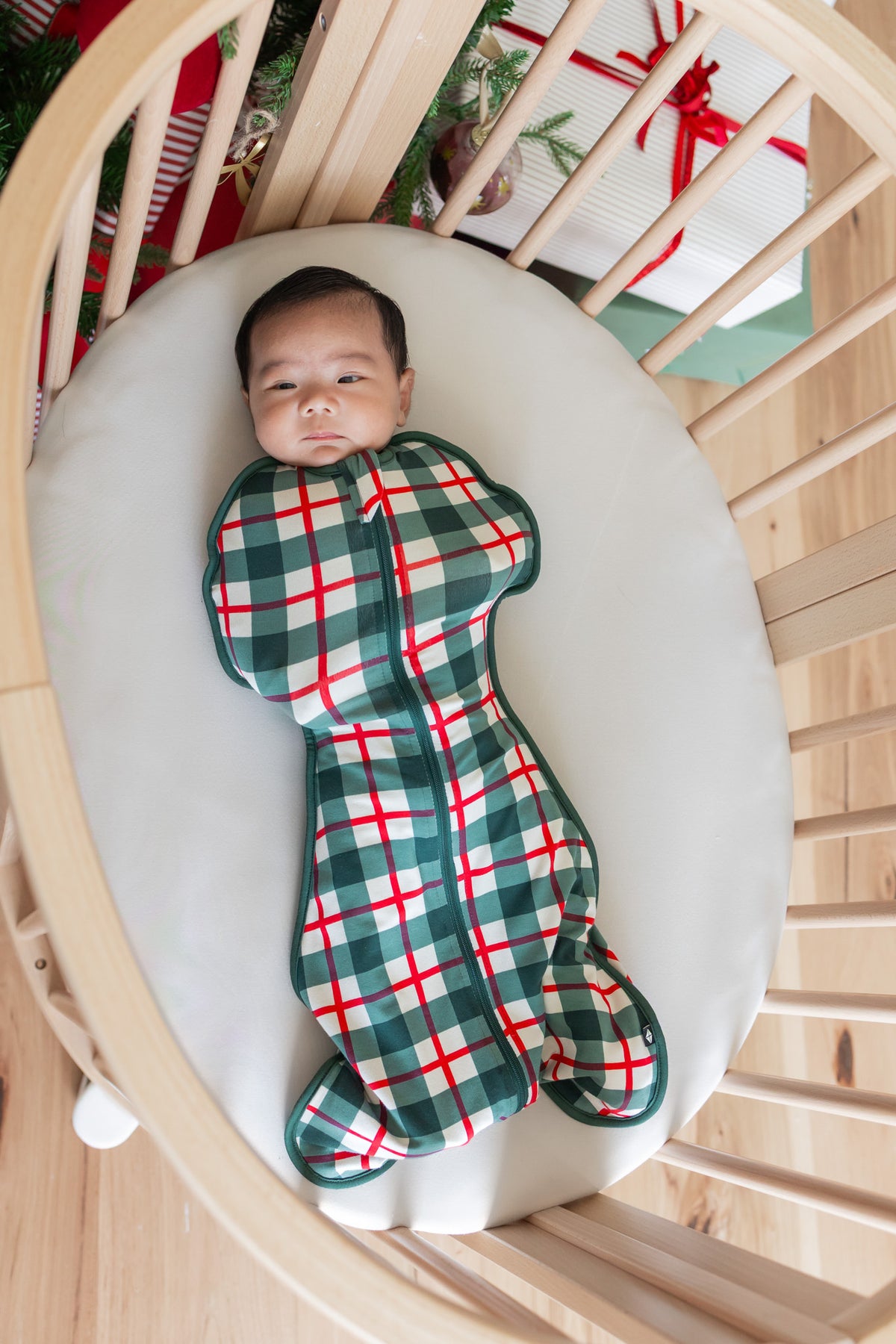 A baby lying on their back inside a wooden crib, swaddled in a green and red plaid swaddle bag