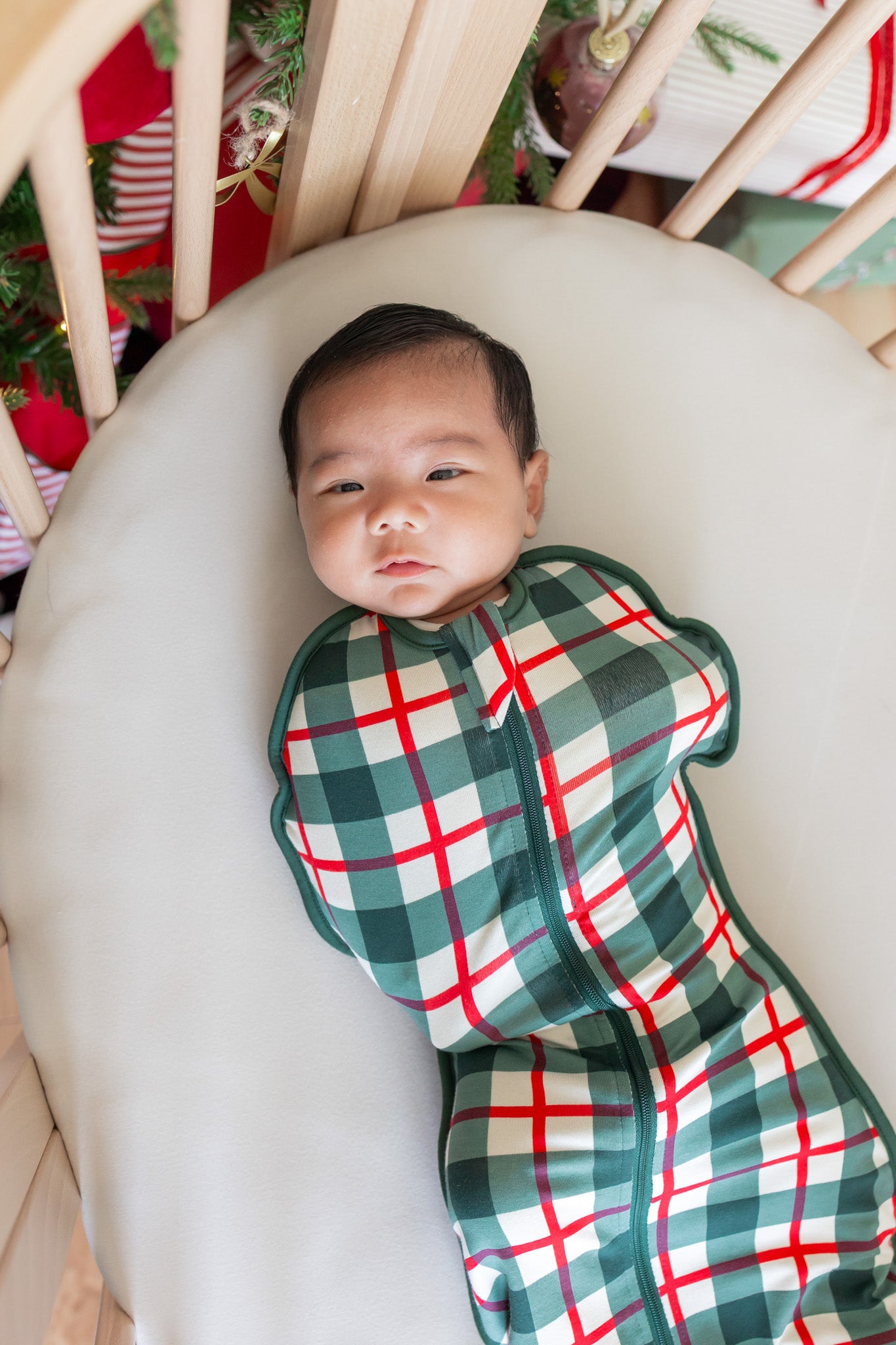 A baby swaddled in a green, white, and red plaid swaddle bag, lying on a white crescent pillow inside a wooden crib.