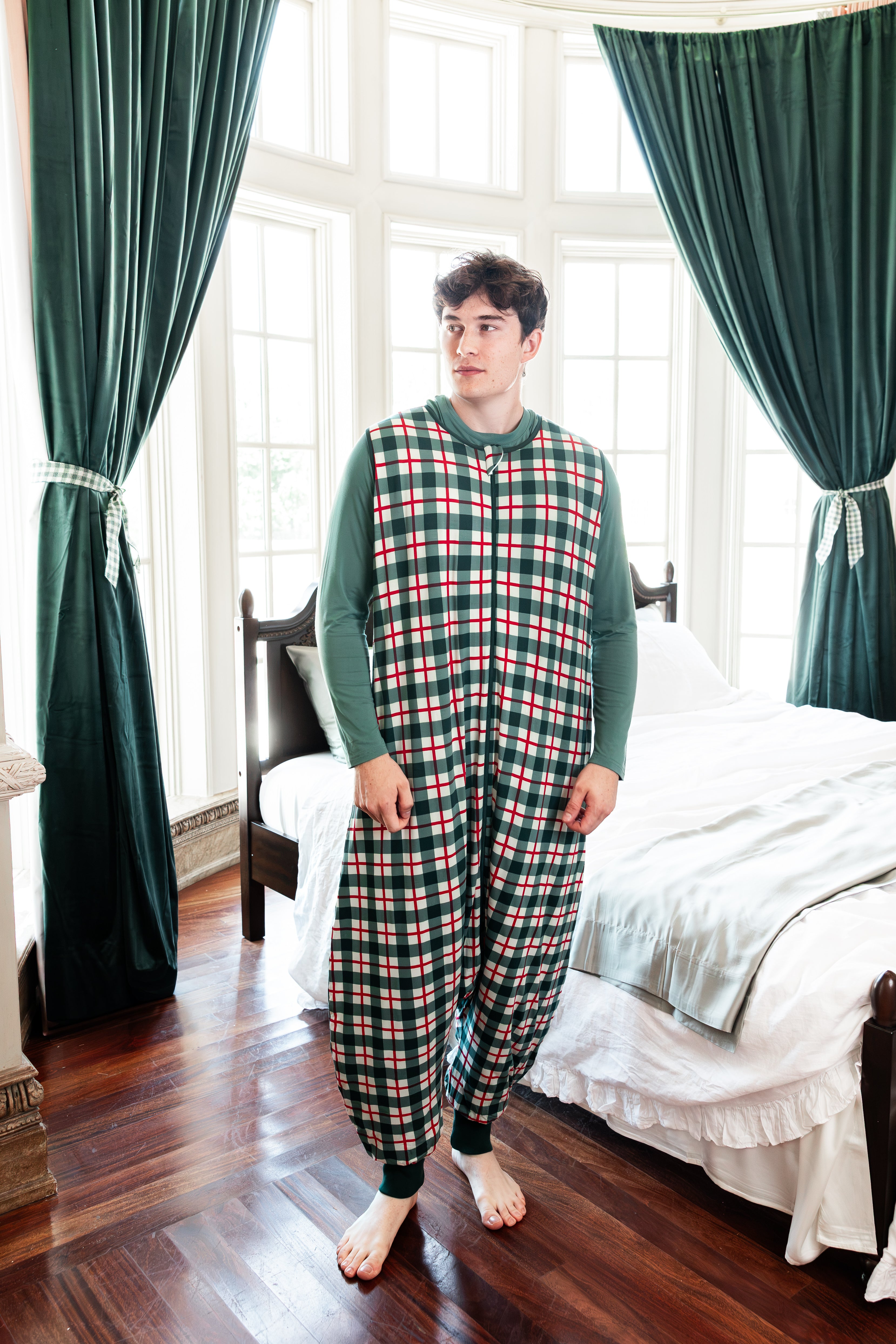 A man wearing a green, red, and white plaid sleep sack with solid green long sleeves, standing in a bedroom with dark green curtains and a bed in the background.