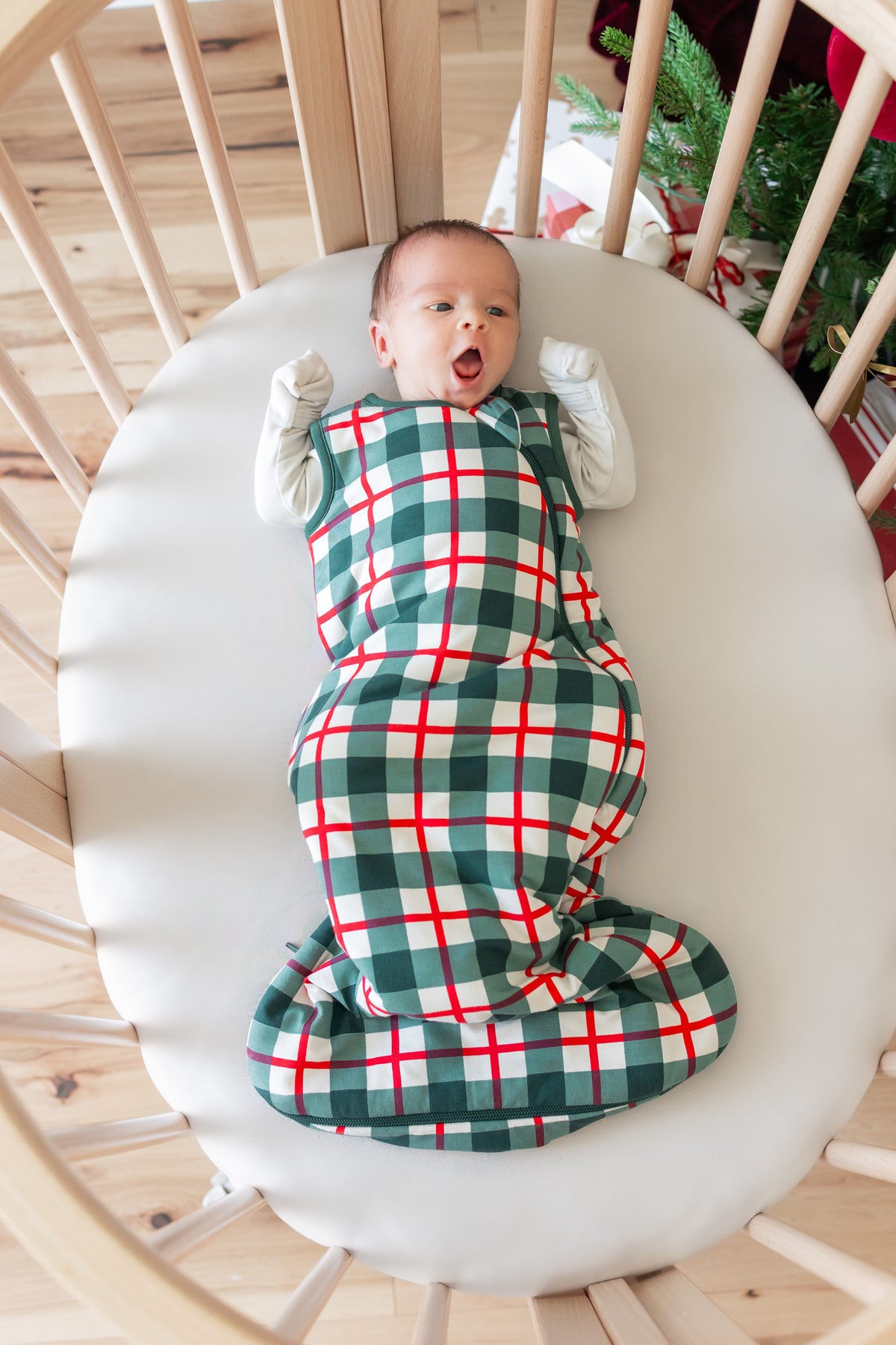 A baby lying on their back inside a wooden crib, wrapped in a green and red plaid sleep sack over a white long-sleeve outfit.