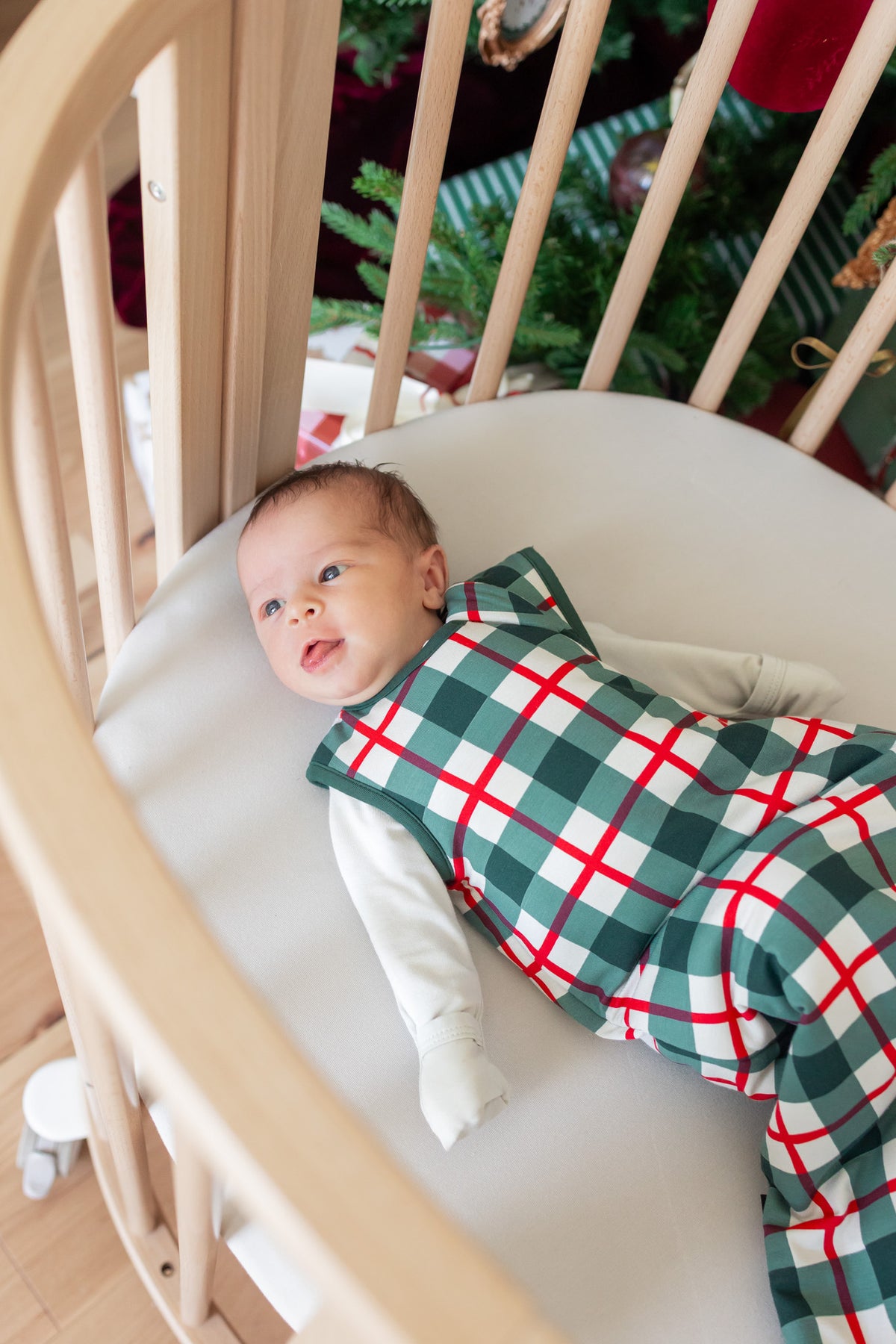 A baby lying in a light wood crib, wearing a green, white, and red plaid holiday sleep sack. A decorated Christmas tree is visible in the background.