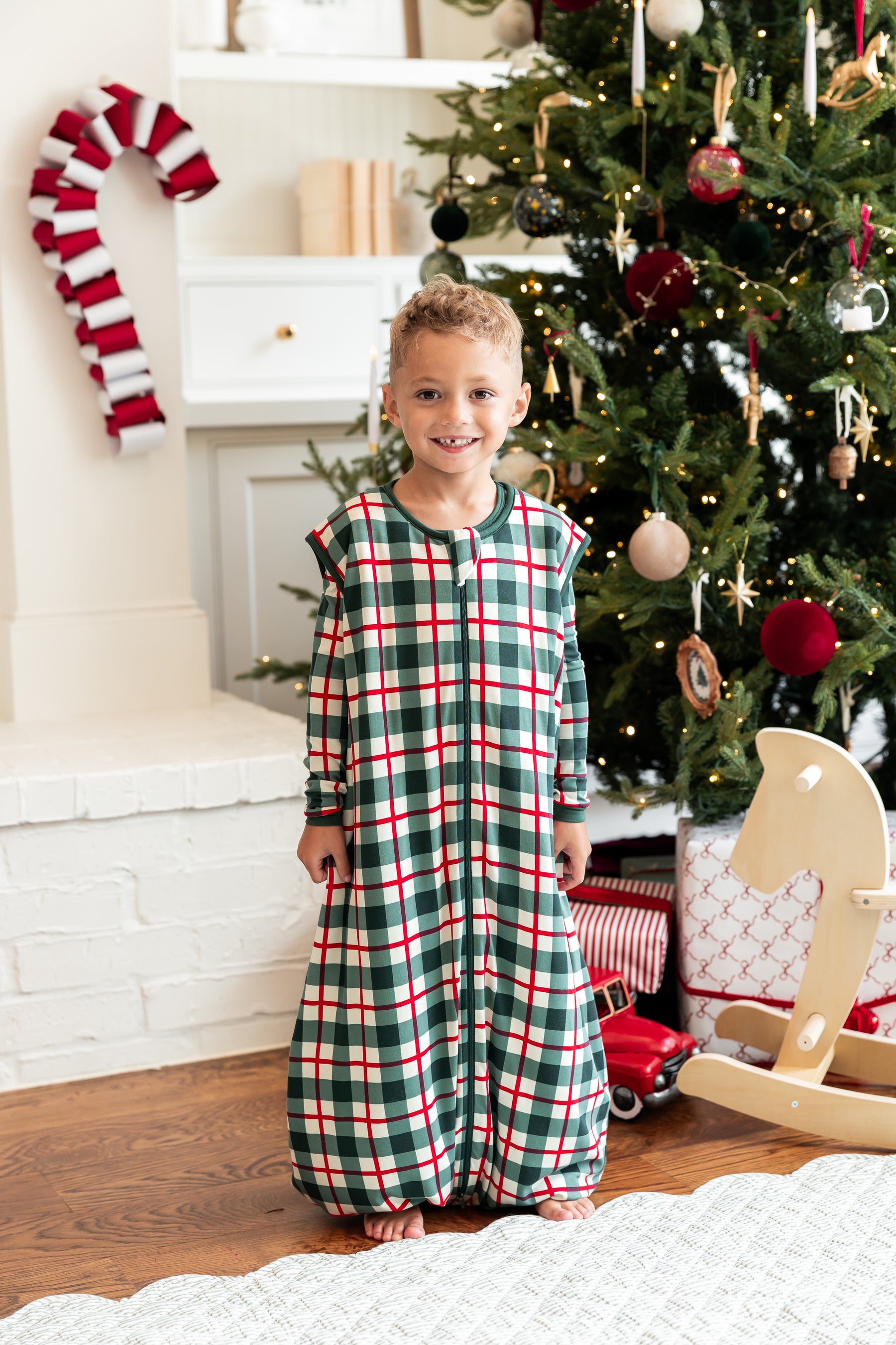A young boy stands barefoot in a living room in front of a decorated Christmas tree, wearing a holiday sleep sack walker with a green, red, and white plaid pattern.