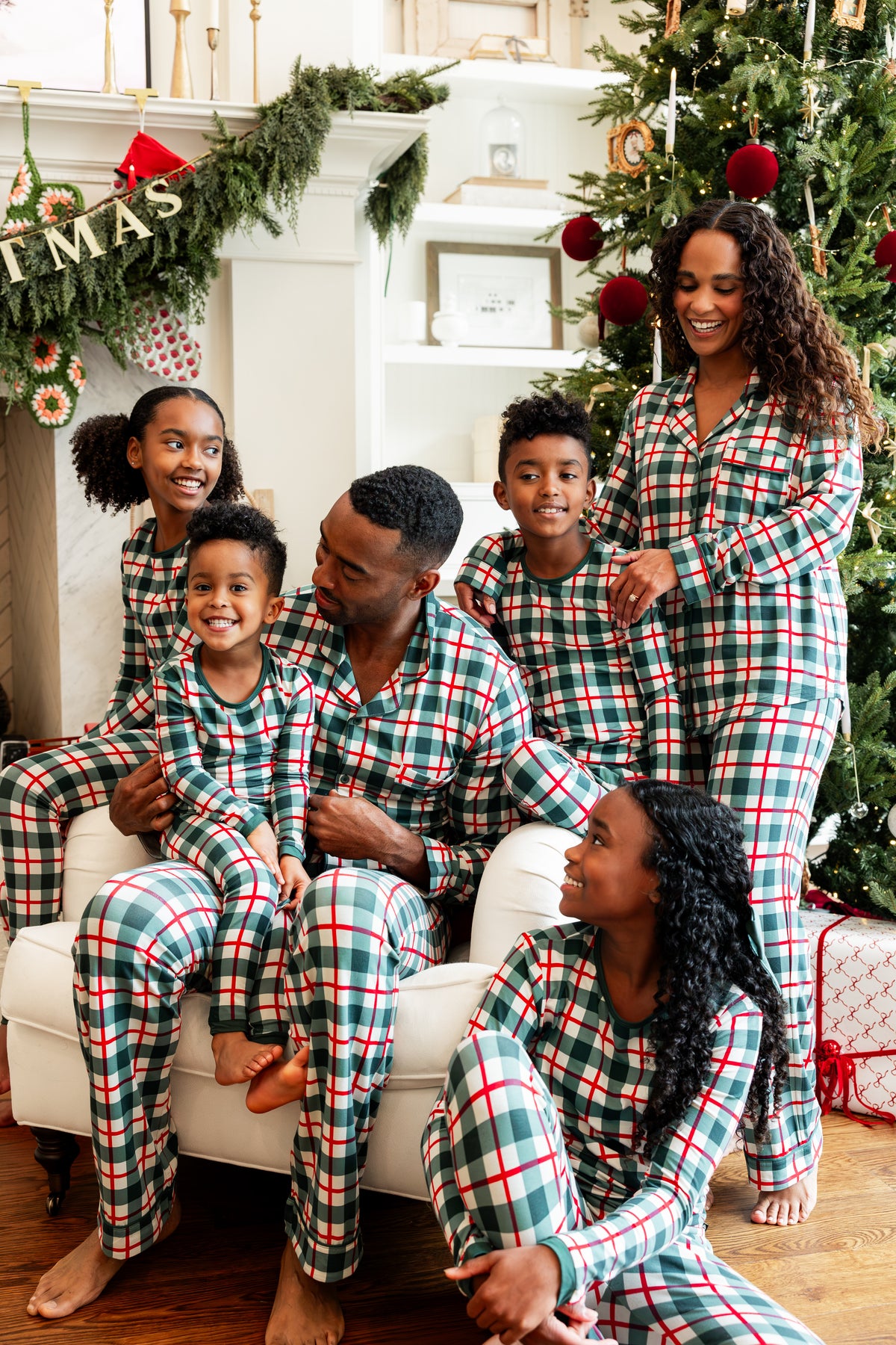 A family of six wearing matching green, red, and white plaid Christmas pajamas, posing together on a white sofa in a decorated living room with a Christmas tree and fireplace.