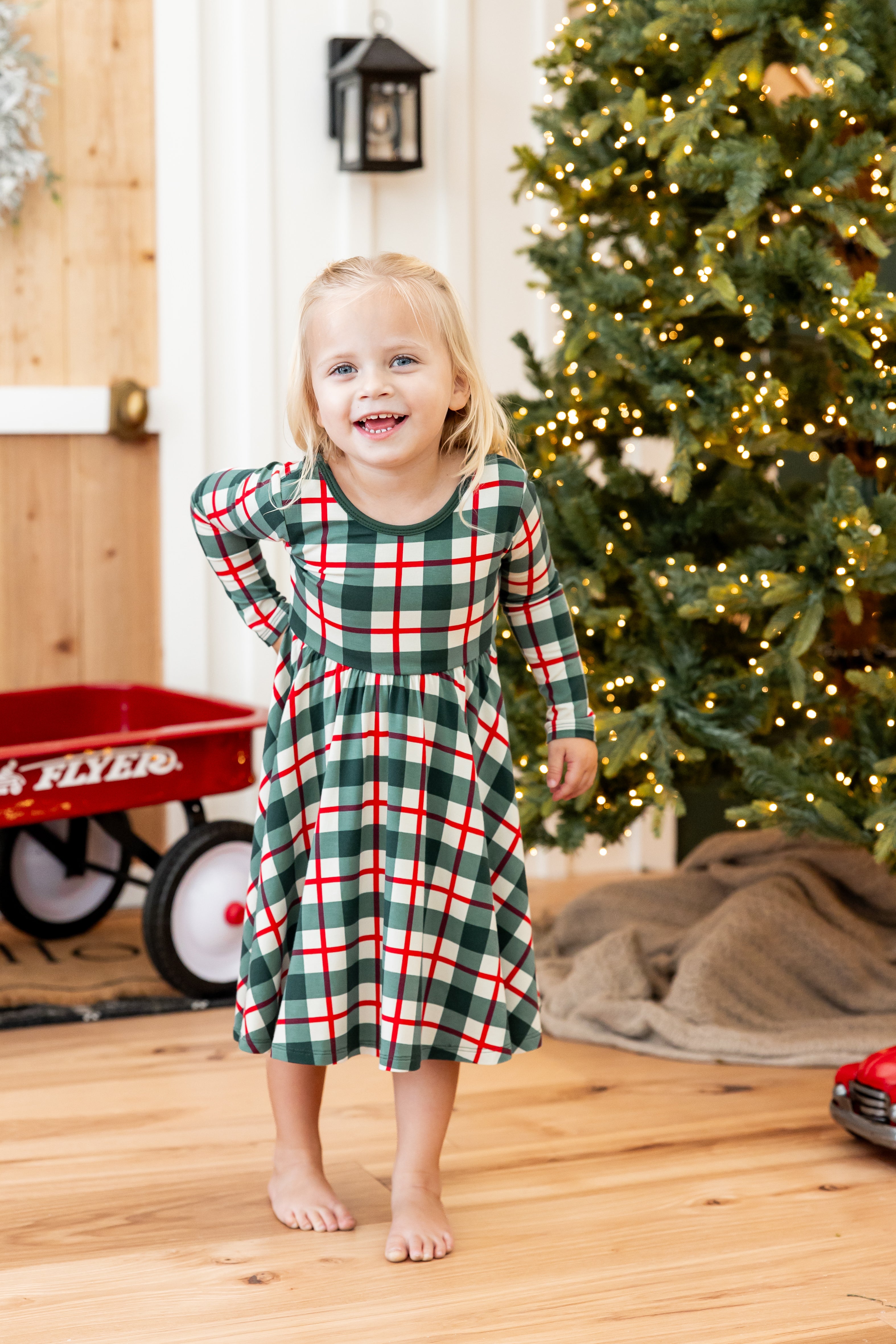 A young girl stands barefoot in a holiday-themed room, wearing a long-sleeve holiday plaid print dress with green, red, and white checkers. A decorated Christmas tree, a red wagon, and a wooden floor are visible in the background.