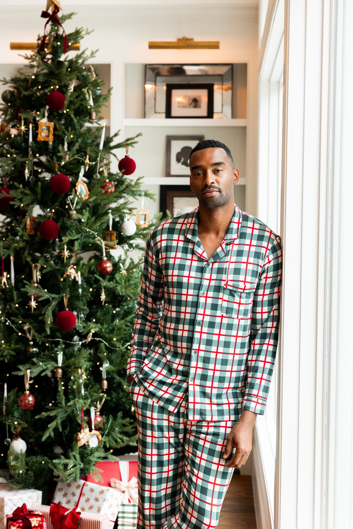 A man wearing a green, red, and white plaid pajama set stands indoors next to a decorated Christmas tree with wrapped gifts and holiday decorations.