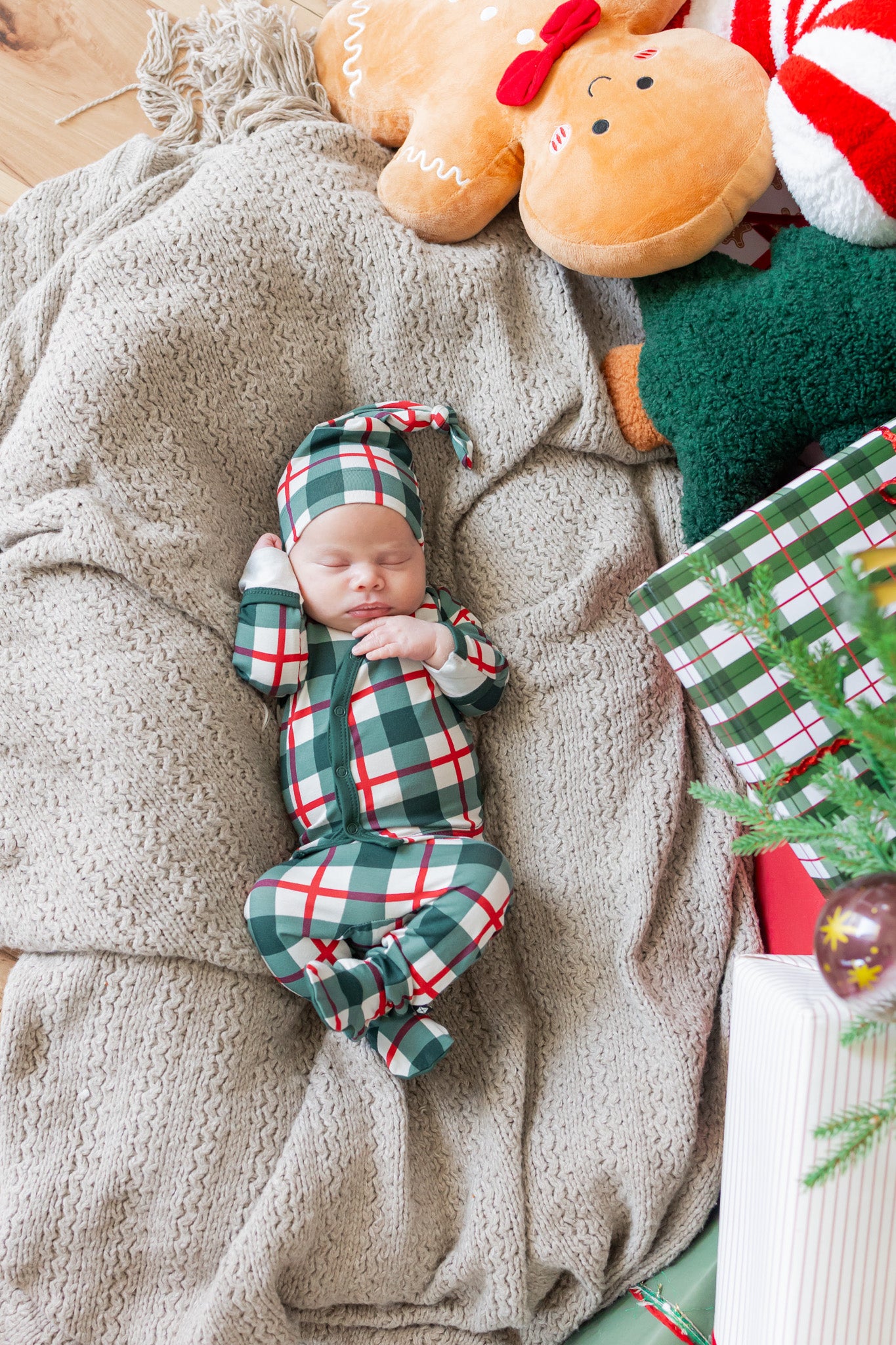 A newborn baby wearing a green and red plaid holiday kimono set with a matching hat, sleeping on a textured beige blanket surrounded by Christmas decorations including a teddy bear, wrapped gifts, and a small tree.