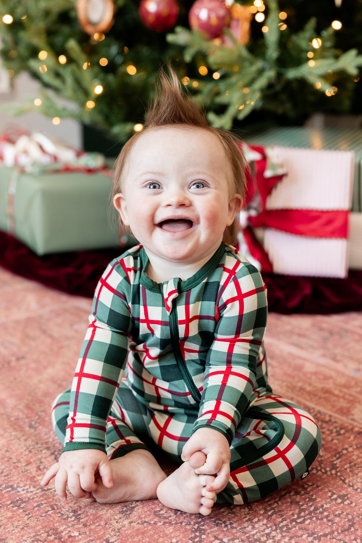 A smiling baby sitting on a patterned floor in front of a decorated Christmas tree and wrapped gifts, wearing a green, red, and white plaid long-sleeve pajama romper.