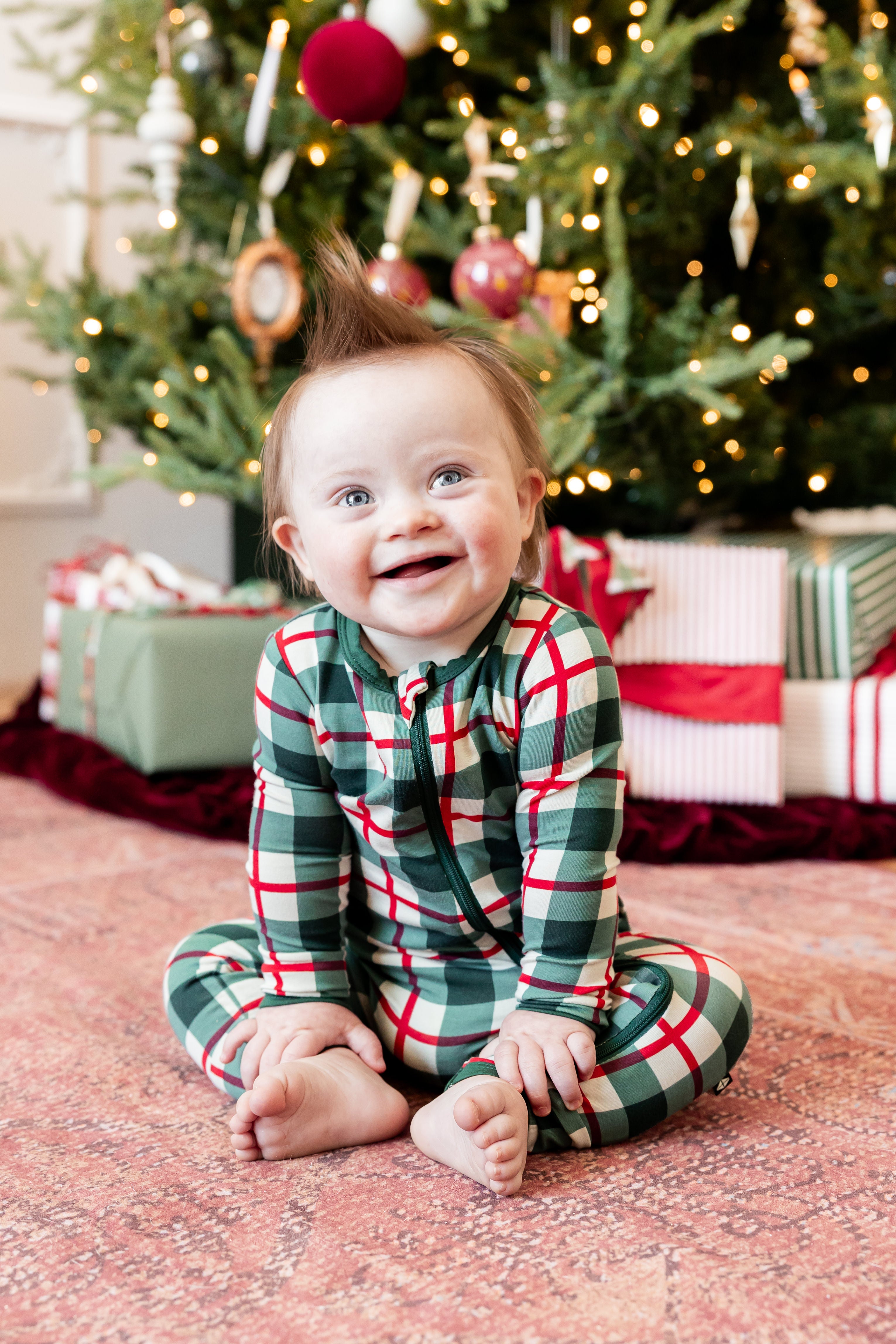 A baby sitting on a patterned rug in front of a decorated Christmas tree, wearing a green, red, and white plaid long-sleeve pajama set.