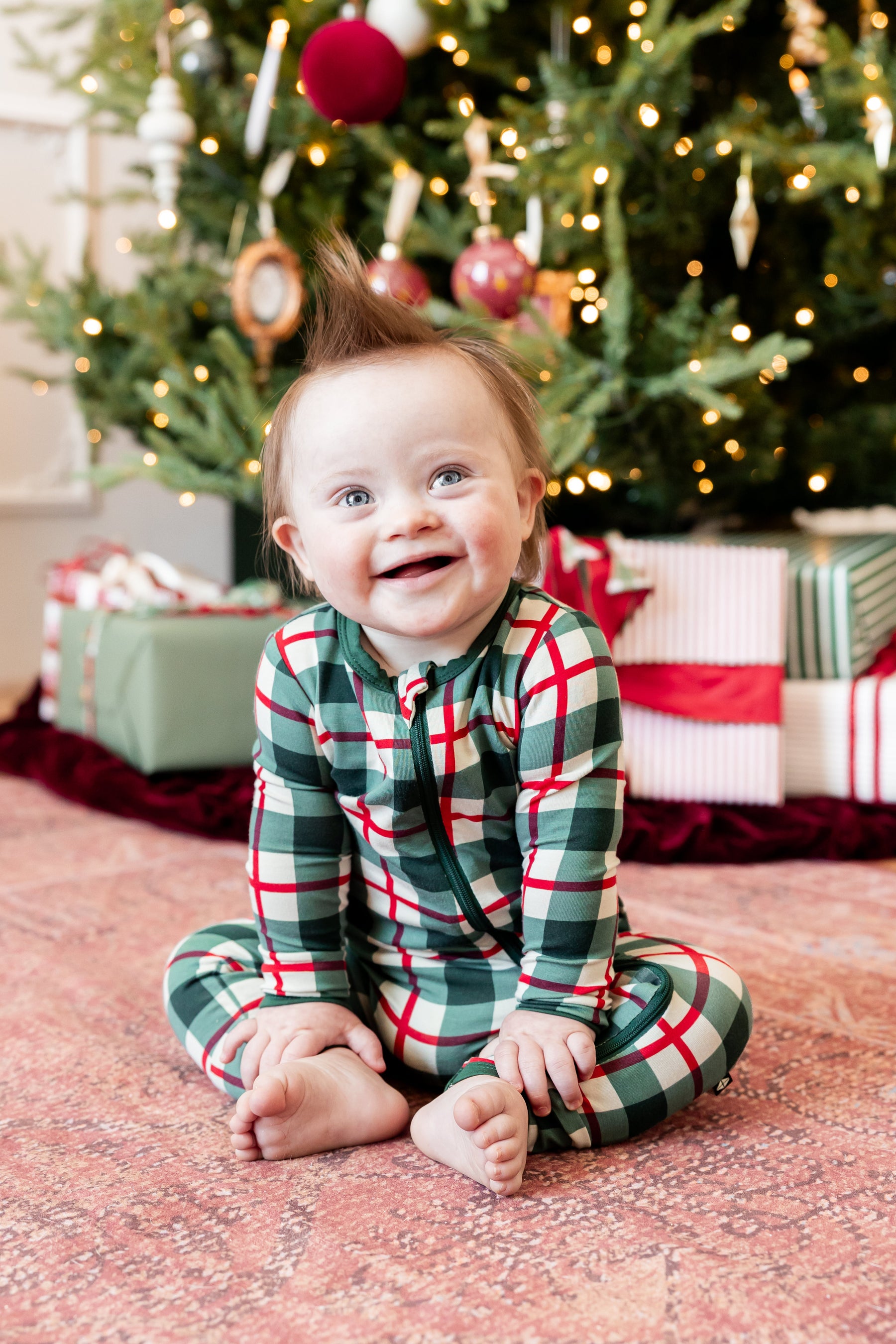 A baby sitting on a patterned rug in front of a decorated Christmas tree, wearing a green, red, and white plaid long-sleeve pajama set.
