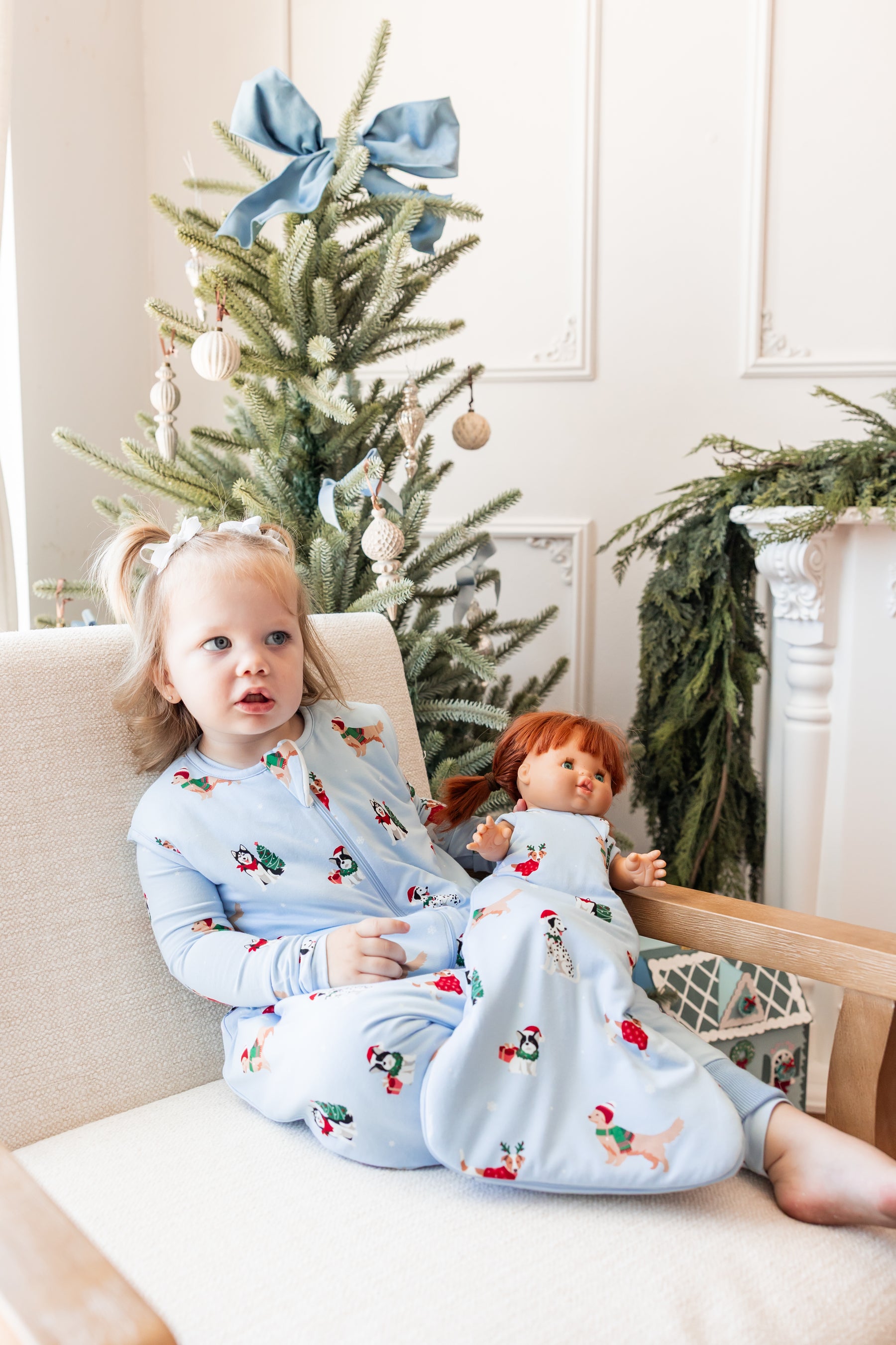 Girl sitting on sofa wearing Sleep Bag Walker in Holiday Pups 1.0 print holding a doll wearing a similar print baby doll sleep bag
