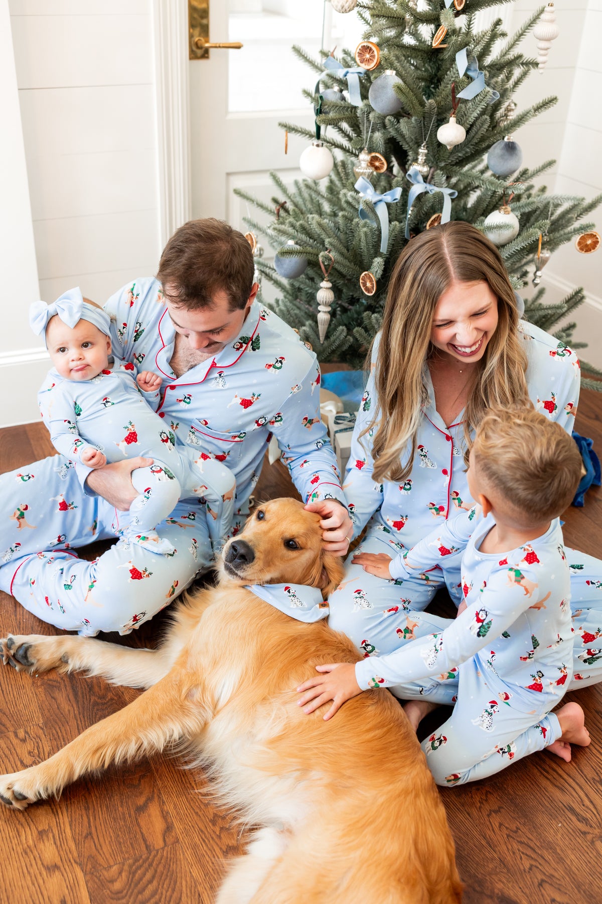 A family of four wearing matching light blue Christmas-themed pajamas sits on a wooden floor in front of a decorated Christmas tree, with a golden retriever lying in the foreground.