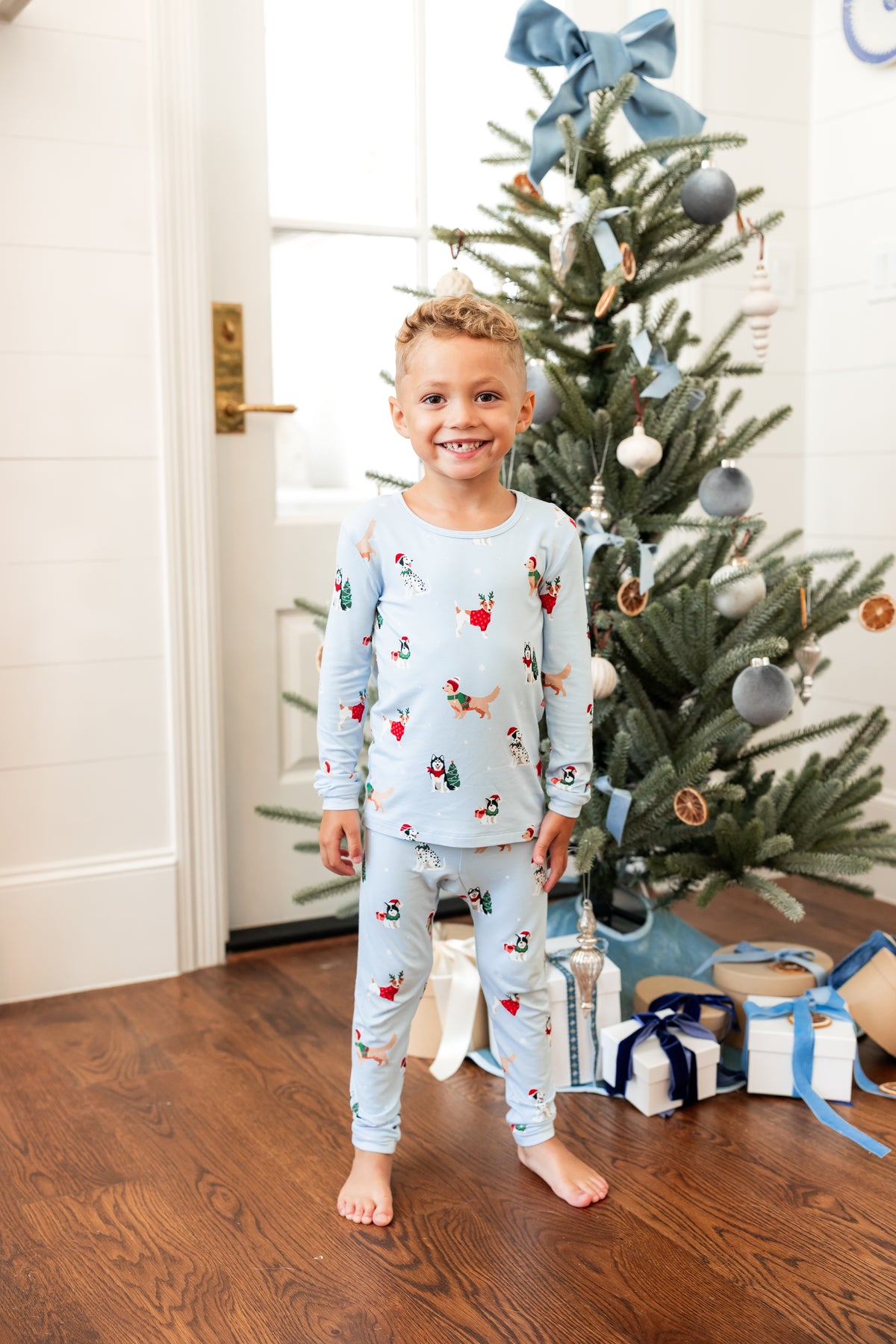 Boy wearing Long Sleeve Pajamas in Holiday Pups print with Christmas tree and presents in the background