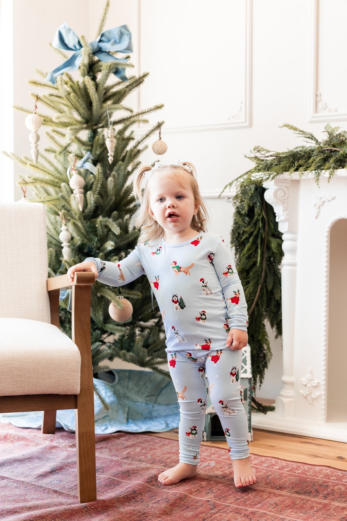 Girl standing beside a chair wearing Long Sleeve Pajamas in Holiday Pups print with Christmas tree in the background