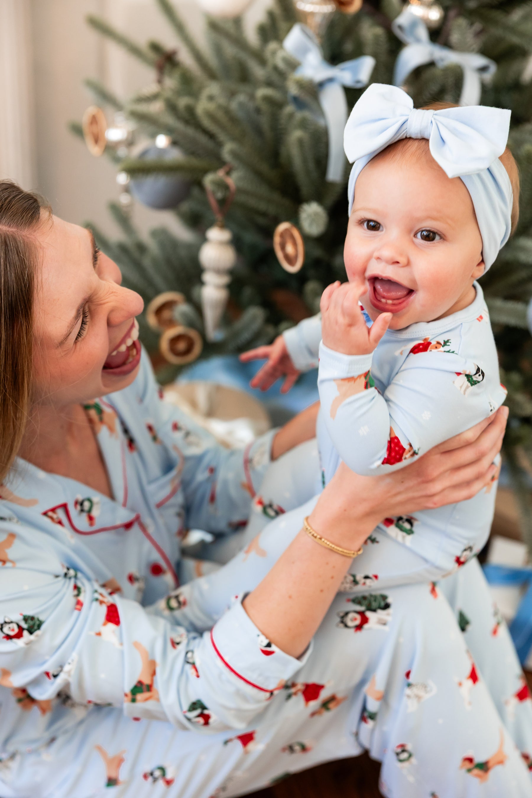 A woman and a baby wearing matching light blue Christmas-themed pajamas sit together in front of a decorated Christmas tree. The pajamas have a festive print with red accents, and the baby also wears a matching light blue bow headband.