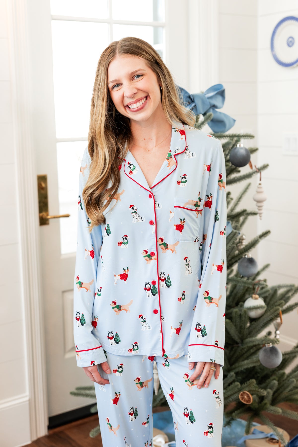 Woman grinning in Long-Sleeved Women's Pajama Set in Holiday Pups print while standing in front of the Christmas tree