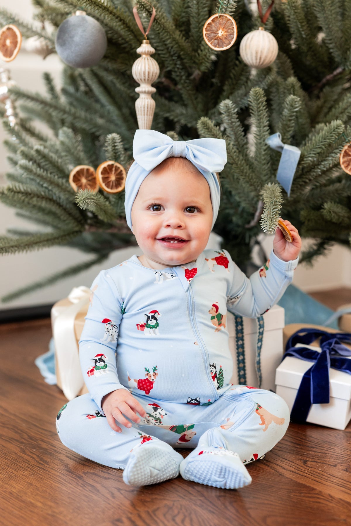 Baby wearing Zippered Footie in Holiday Pups print and blue knotted bow headband with Christmas tree and presents in the background