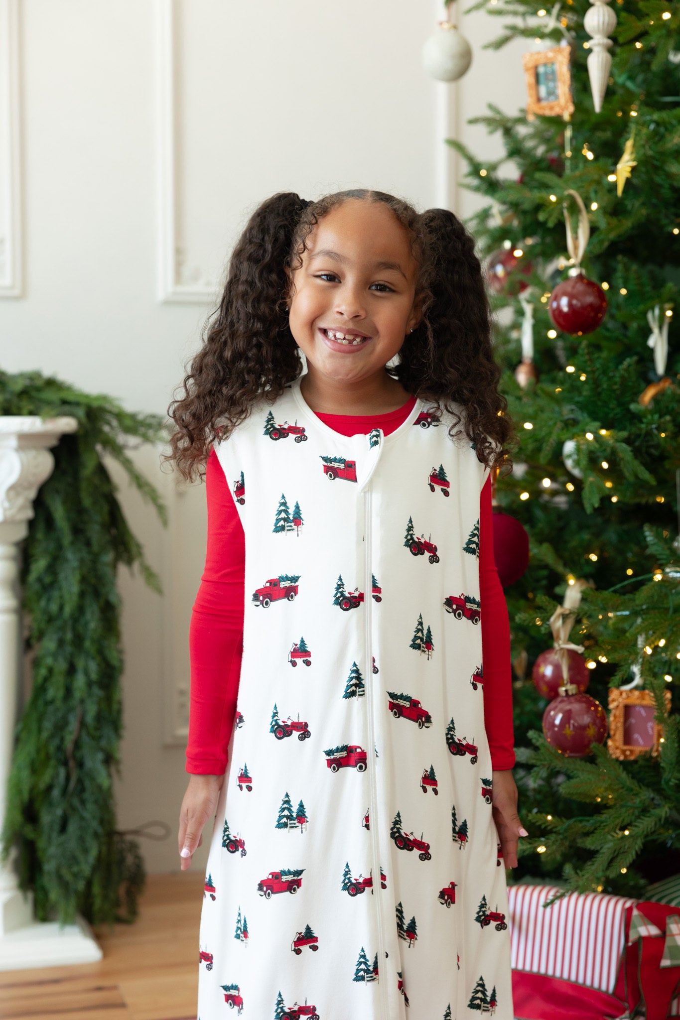 A young girl with curly brown hair in pigtails stands smiling in front of a decorated Christmas tree. She is wearing a white sleeveless holiday sleep sack walker pattern of red trucks and green Christmas trees, layered over a red long-sleeve shirt.