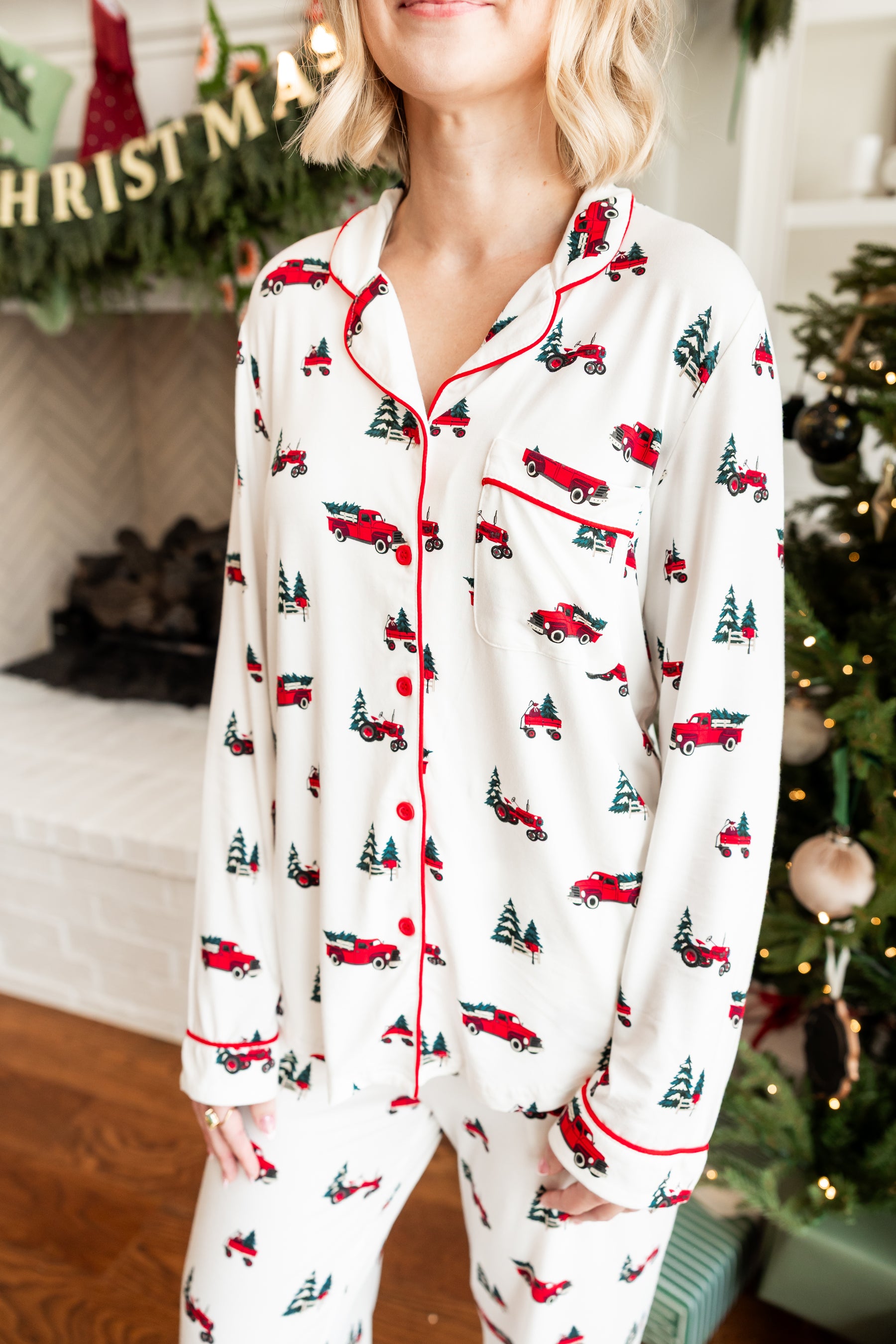 A woman wearing a two-piece Christmas pajama set with an off-white base, red piping, and a repeating pattern of red fire trucks and green Christmas trees, posed in a cozy holiday living room setting.