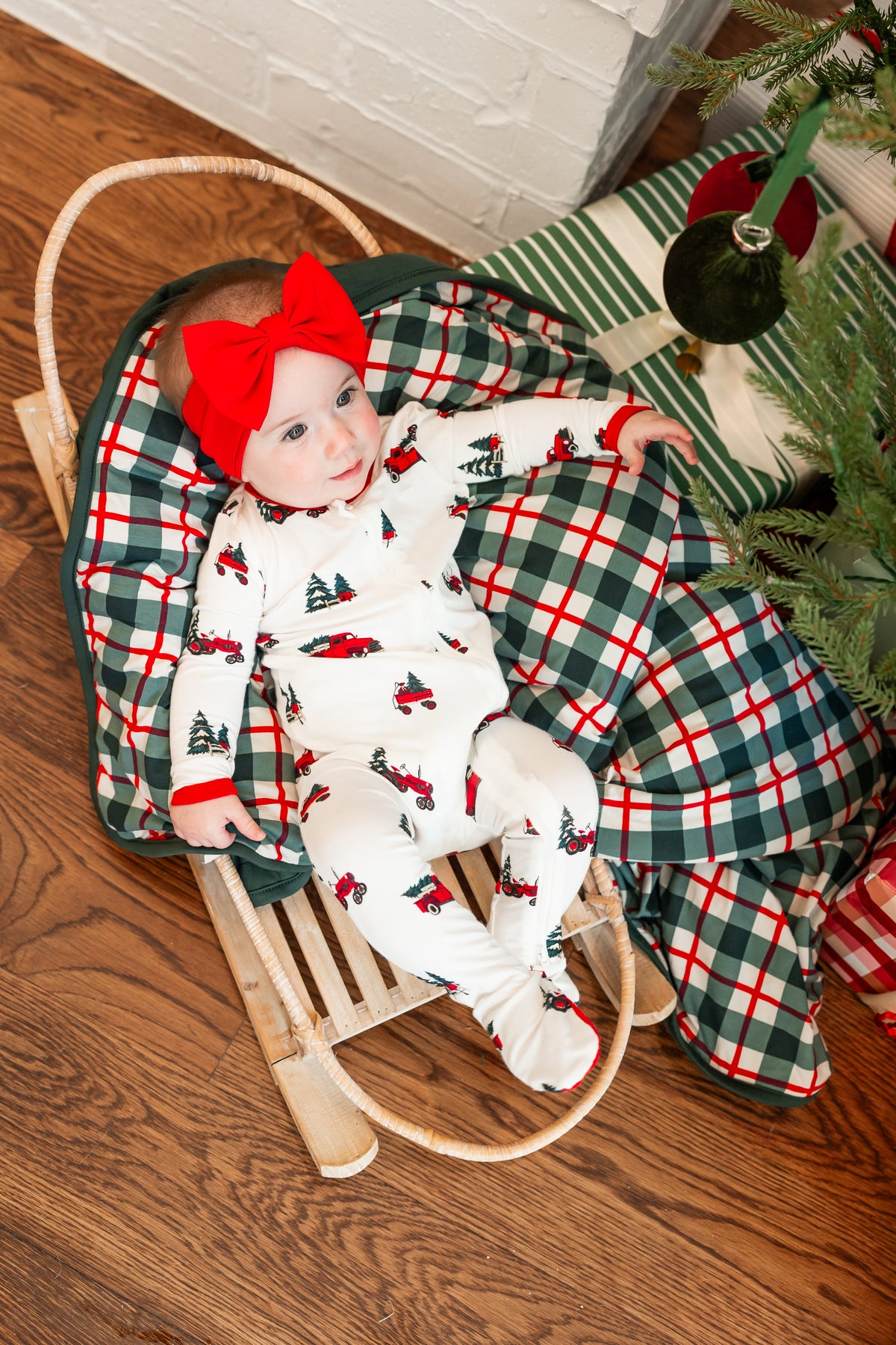 A baby wearing a white footed onesie with a red and green Christmas tree print, a large red bow headband, and a matching plaid blanket, lying in a wooden sled next to a Christmas tree on a hardwood floor.